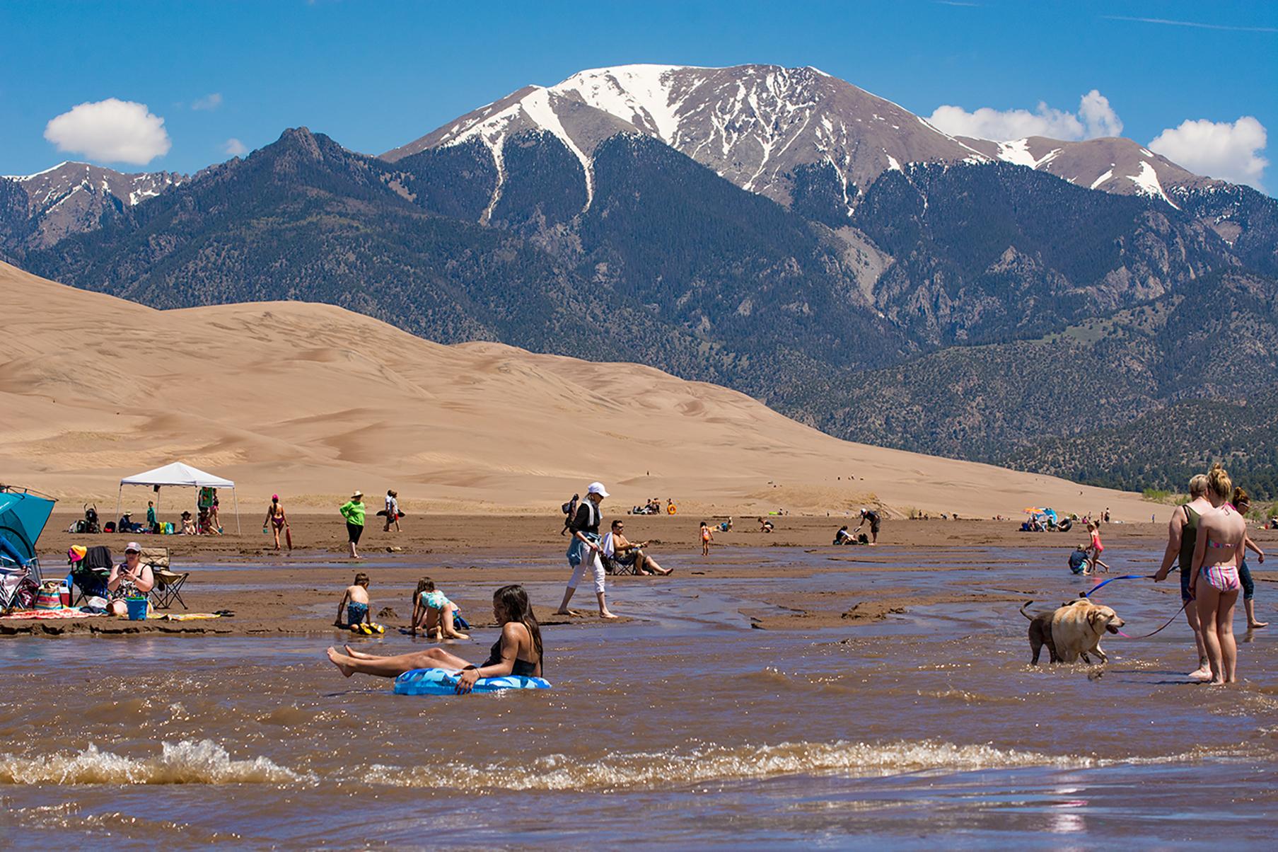 Visitors splashing, floating, and walking in a stream, with dunes and snow-capped mountain.