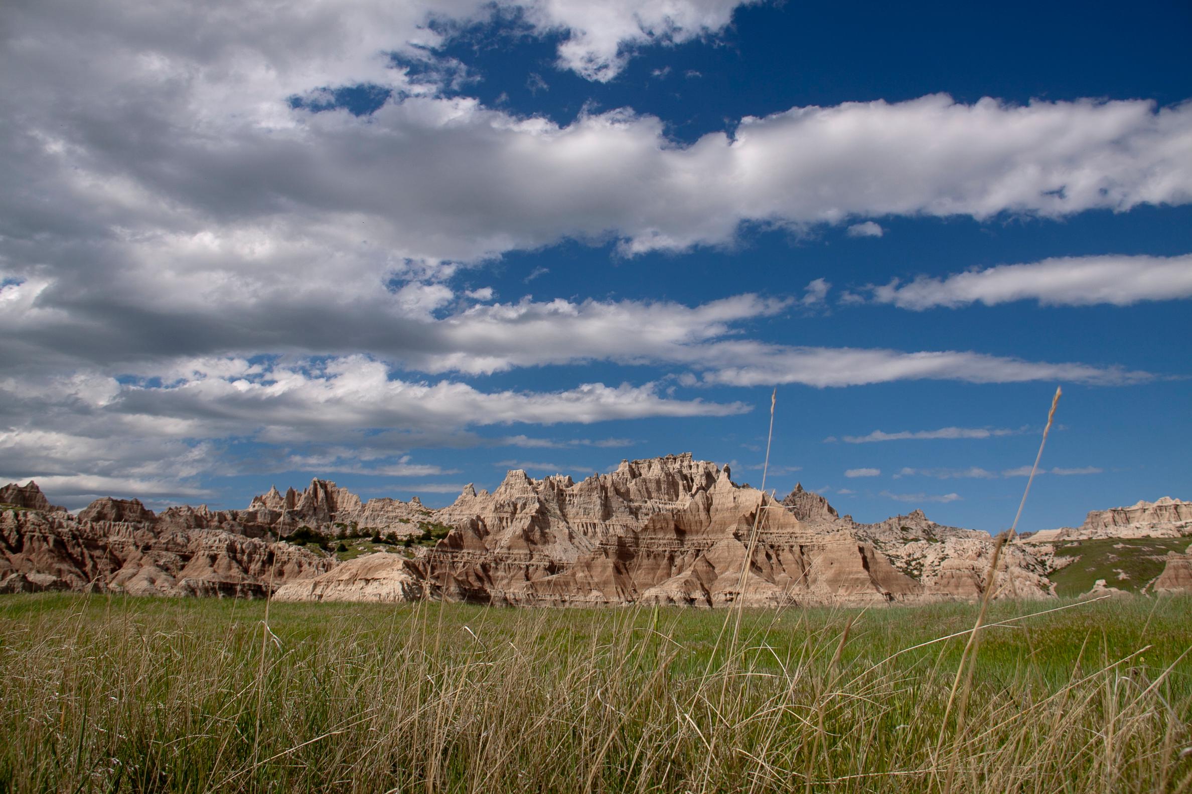 a cloudy blue sky appears over jagged badlands formations, framed by green grasses in the foreground