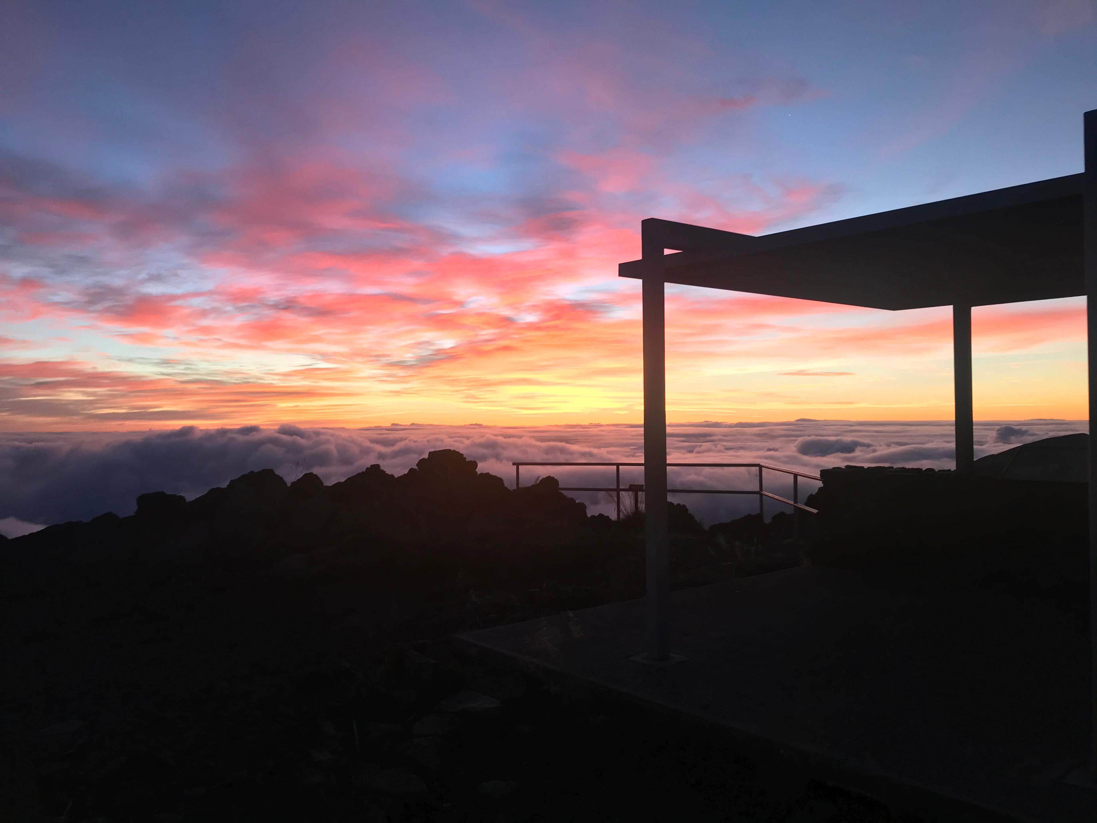 A dawn sky colors clouds yellow and pink, silhouetting an overlook shelter.