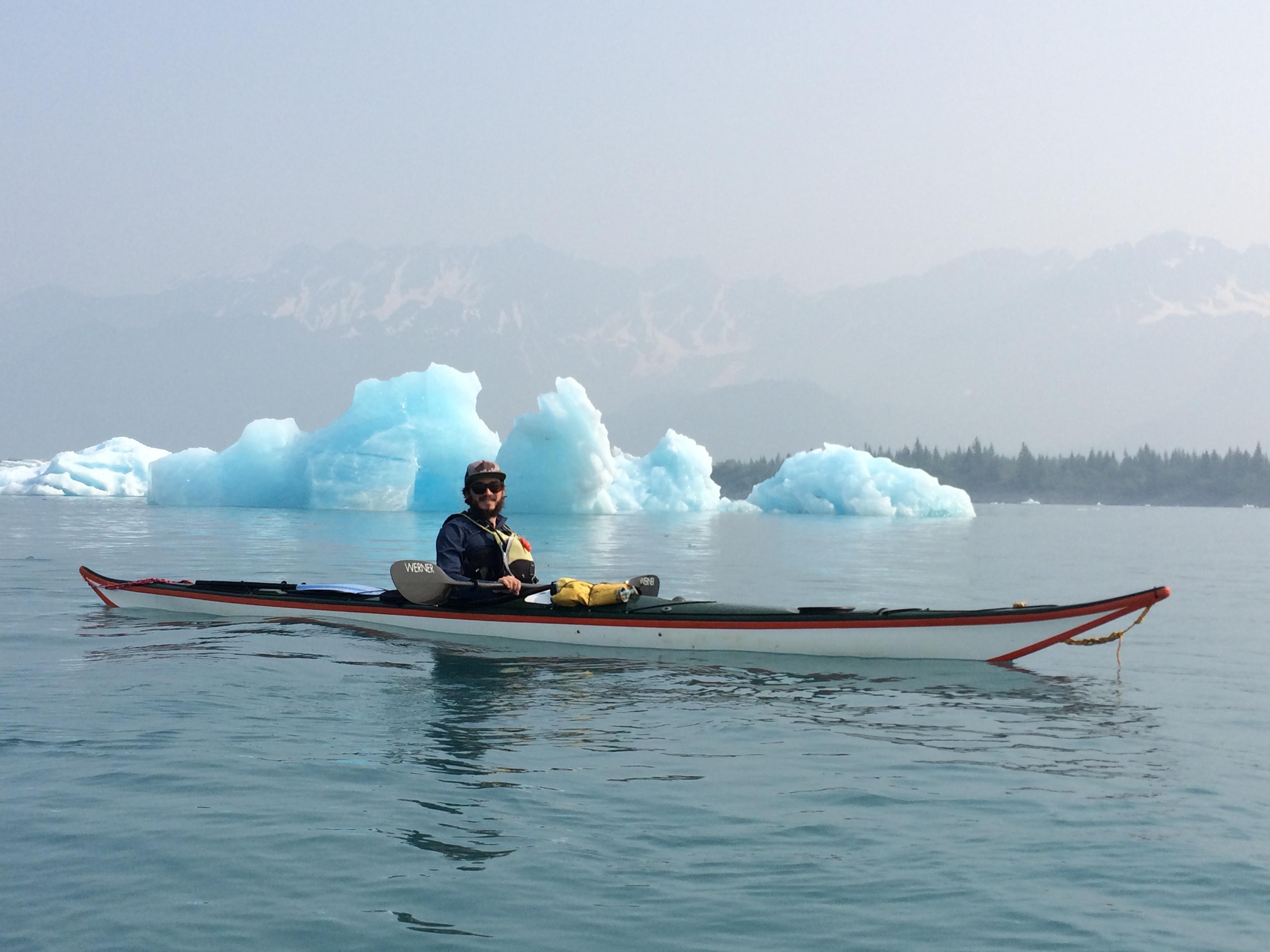 A kayaker is in front of a large blue colored iceberg in the background.