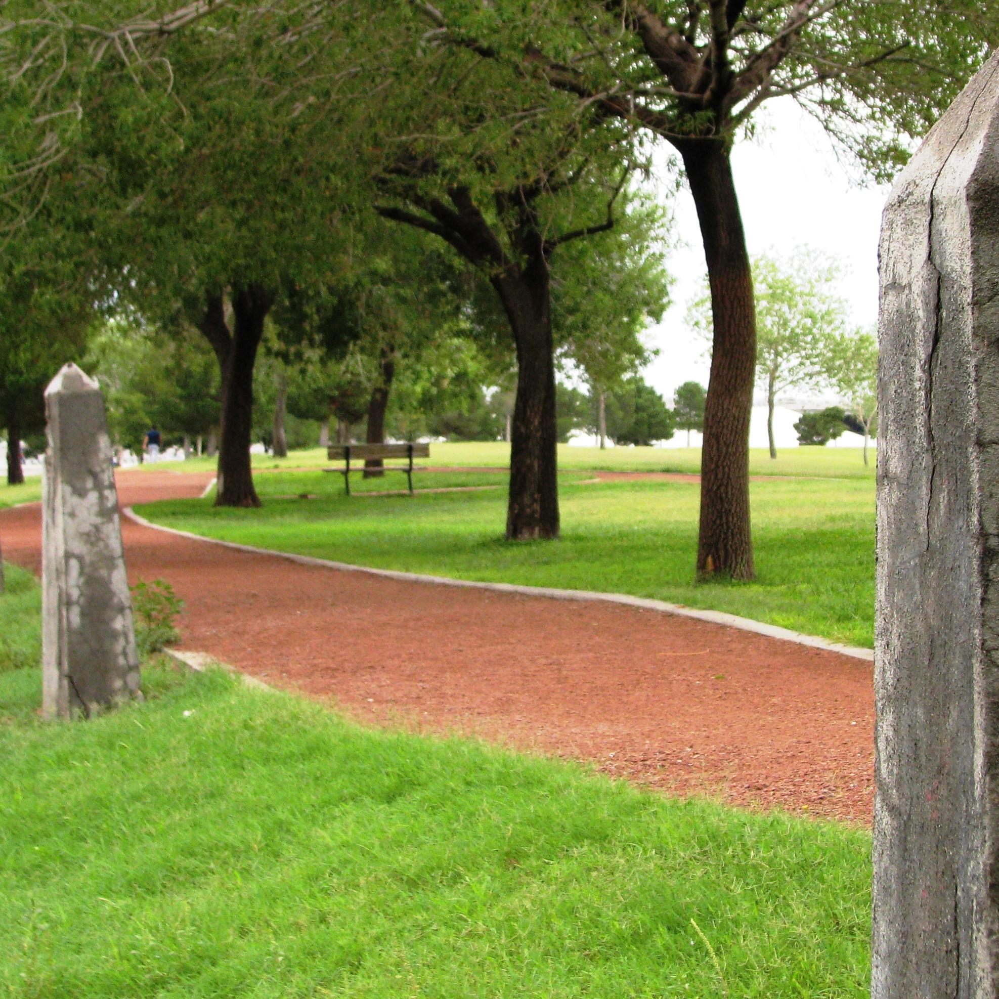 A red gravel trail that has trees and grass surrounding it with 2 boundary fence posts.