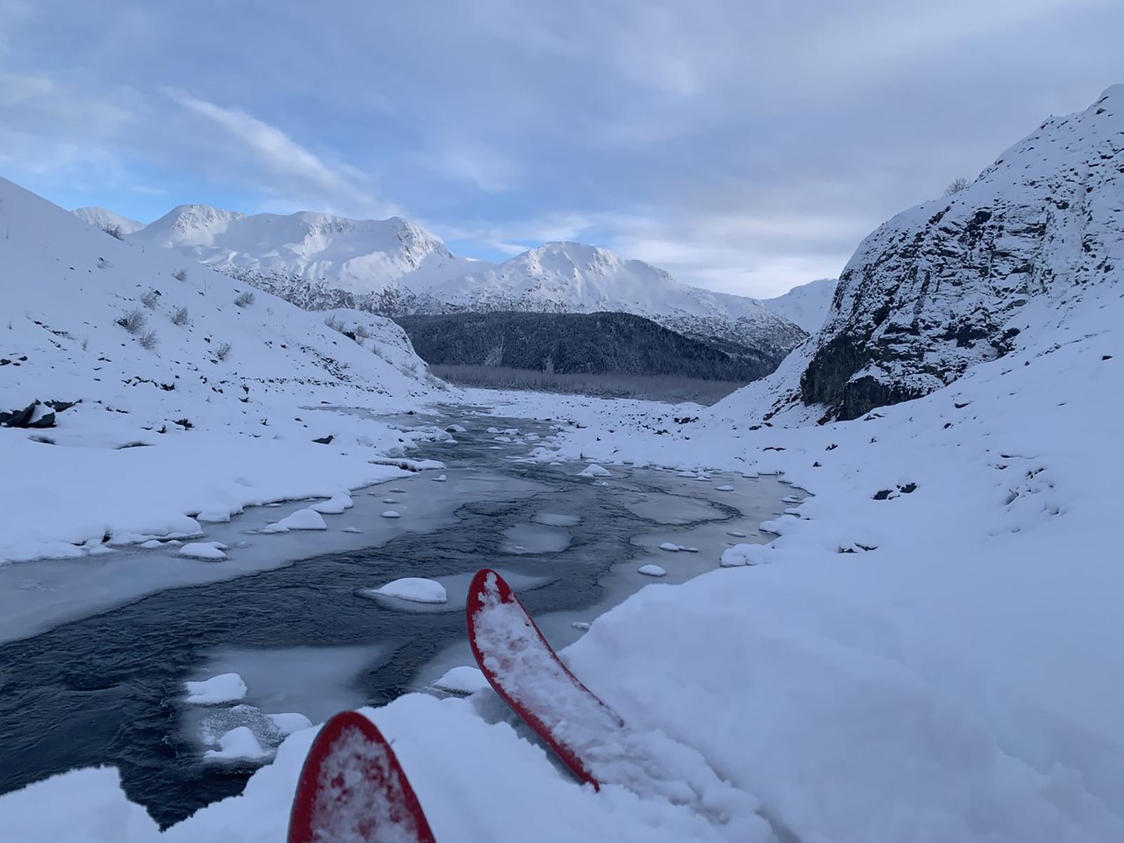 A creek runs through the image center. The land is covered in snow. Ski tips are at the photo bottom