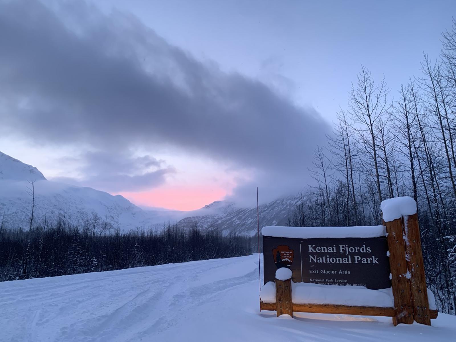 The Kenai Fjords park sign with snow on top. The landscape behind the sign is covered in snow.