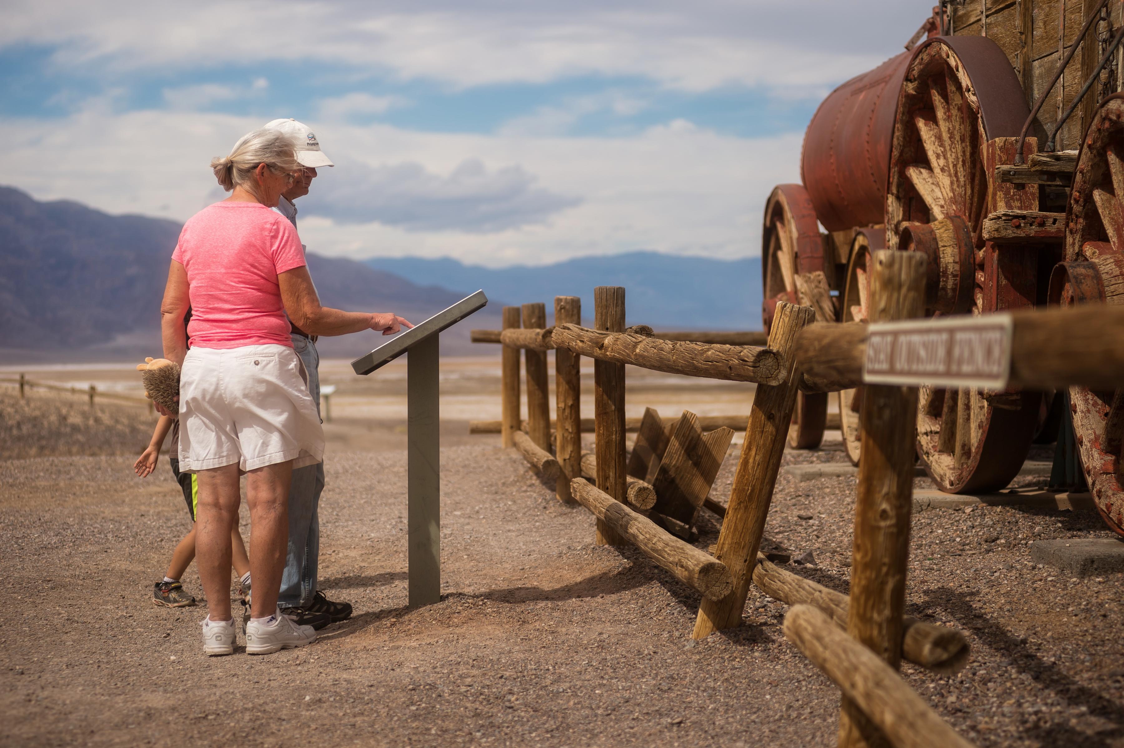 Two adults and a child look at a waist high sign next to a wooden and metal wagon in the desert.