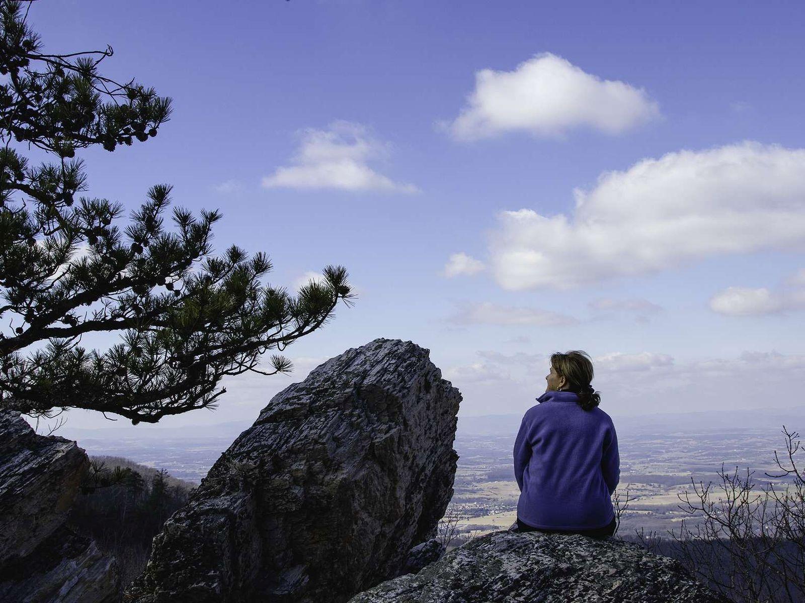 A color photograph of a woman sitting on a rock atop a mountain looking to the side.