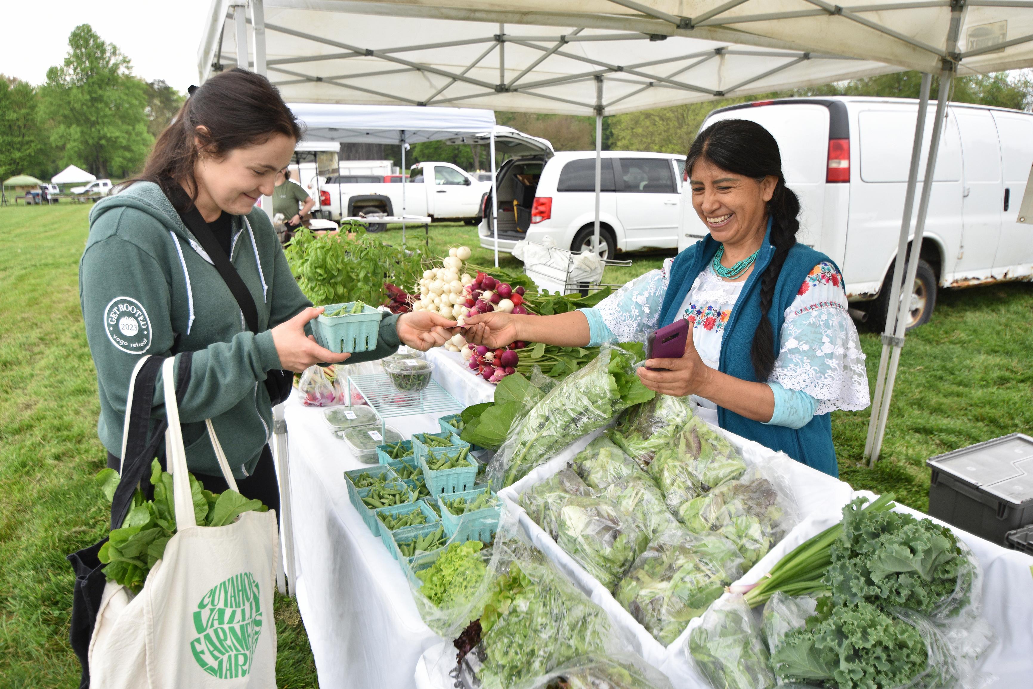 A shopper reaches over table of vegetables to pay a smiling female vendor in embroidered shirt.