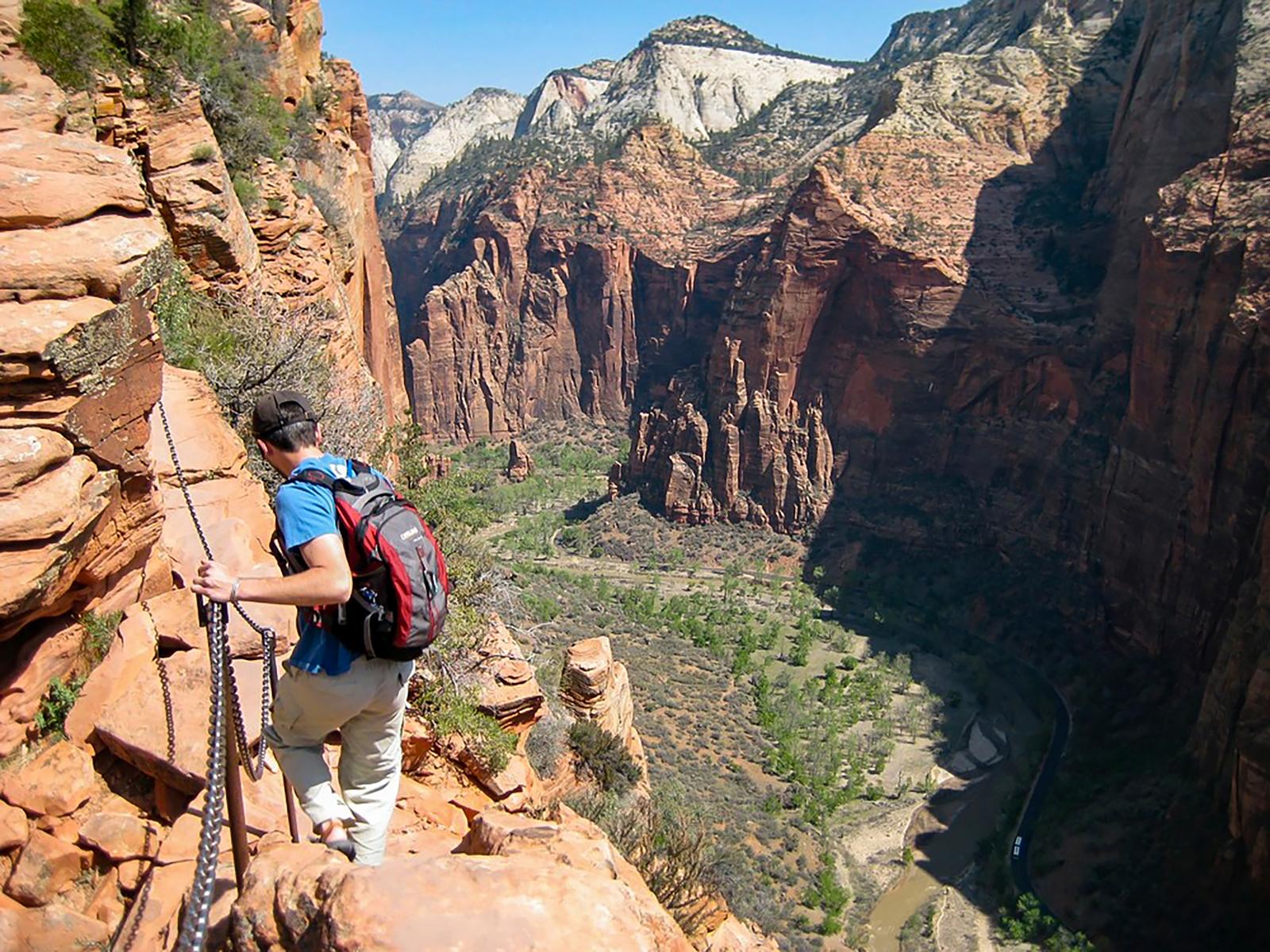 A hiker in a blue shirt holds onto chains as they traverse a sandstone cliff.