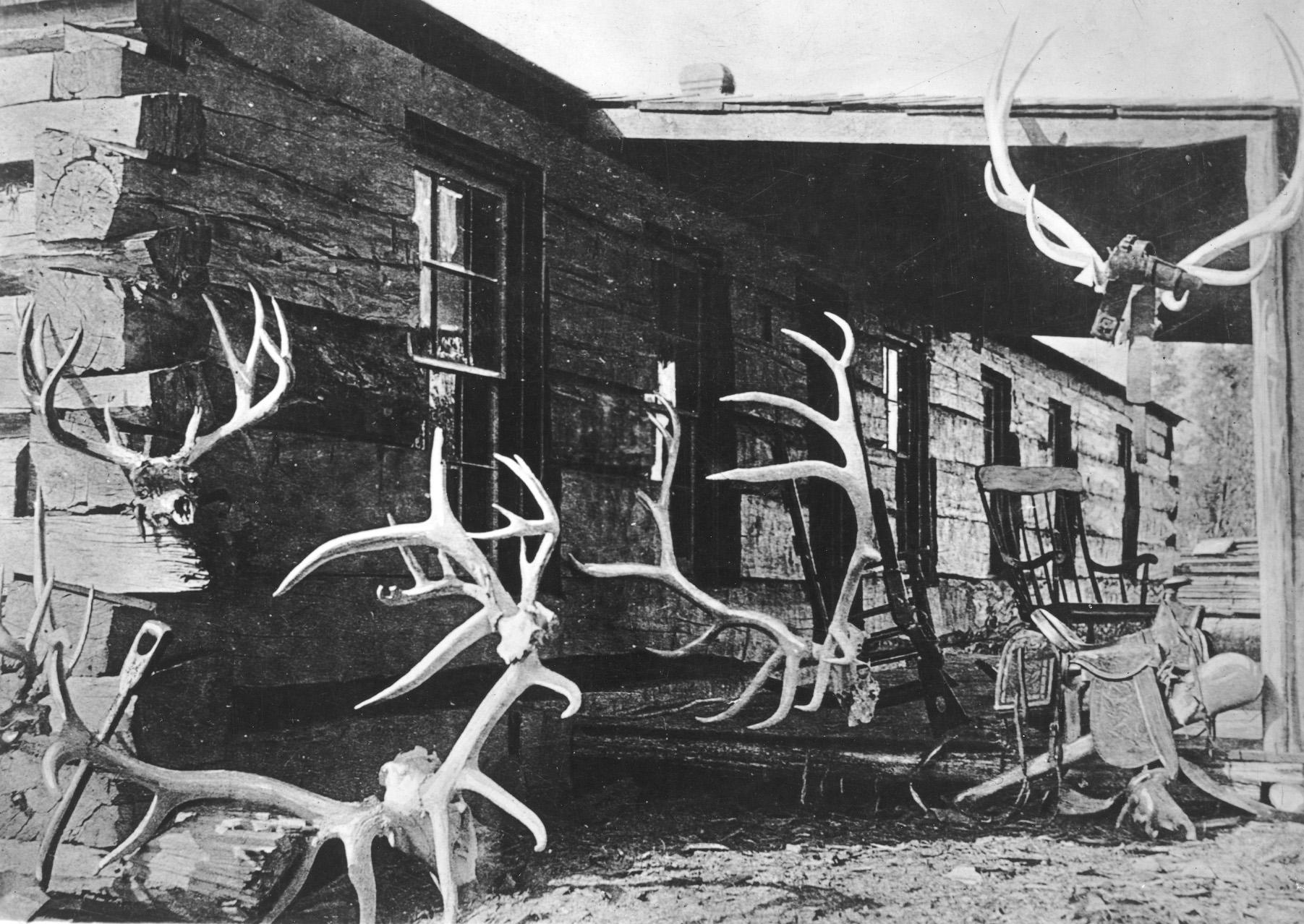 A black and white photo of a low ranch house, with a porch. Elk antlers hang on the walls.