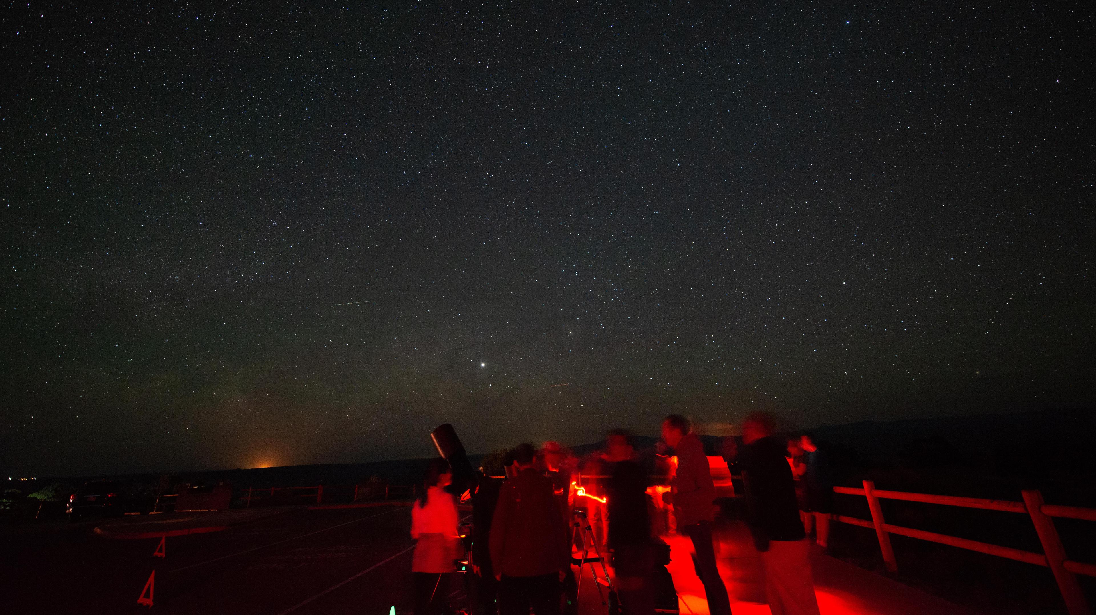 people illuminated in red gather around a telescope pointed at a starry sky