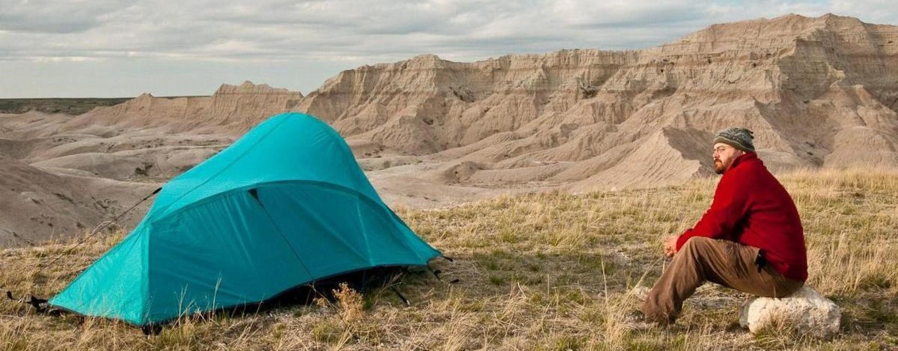 a hiker in a red jacket sits next to a blue tent with badlands buttes in the background