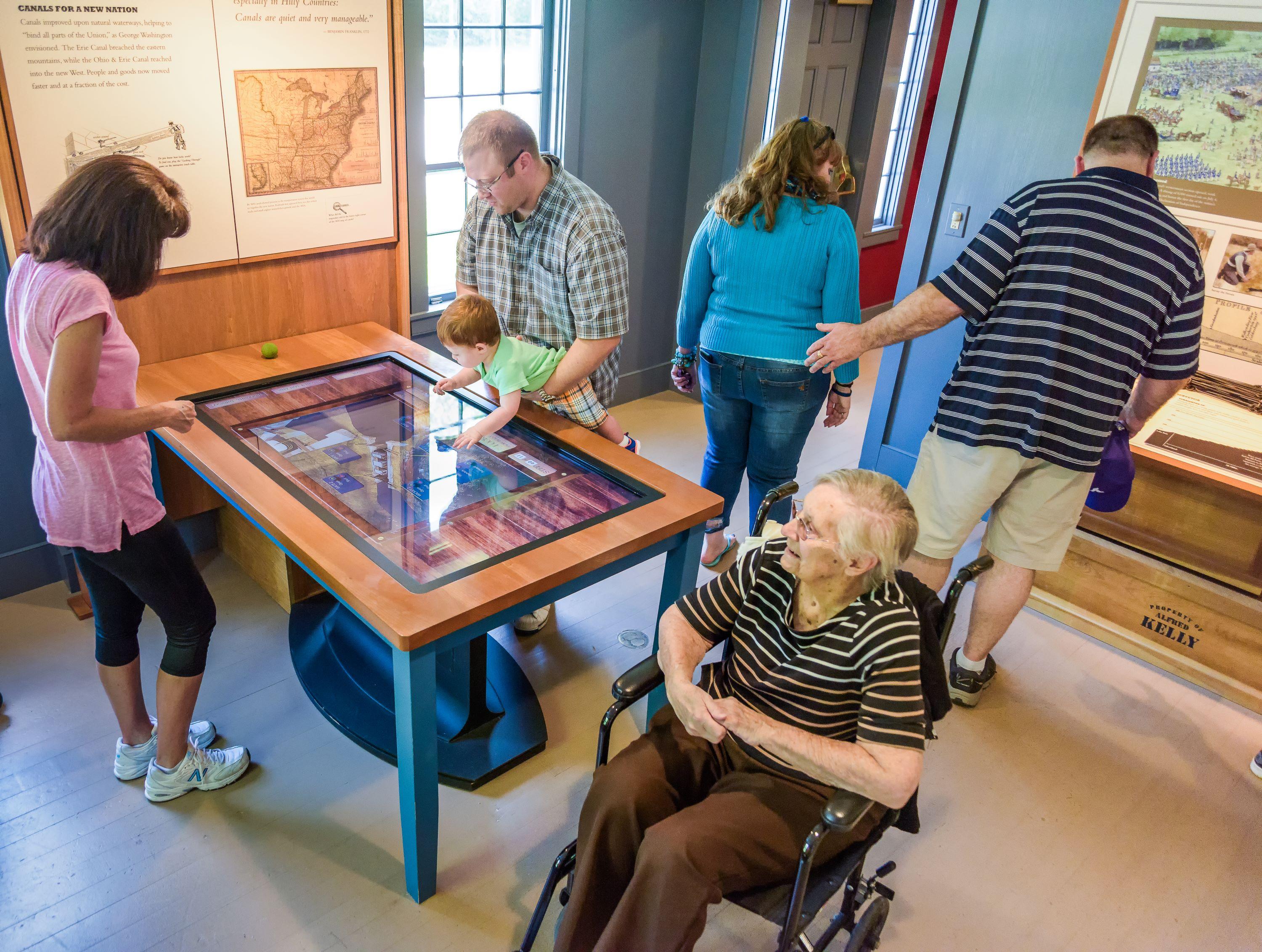 Man holds a child over a touch table as four other adults look at exhibits; one uses a wheelchair.