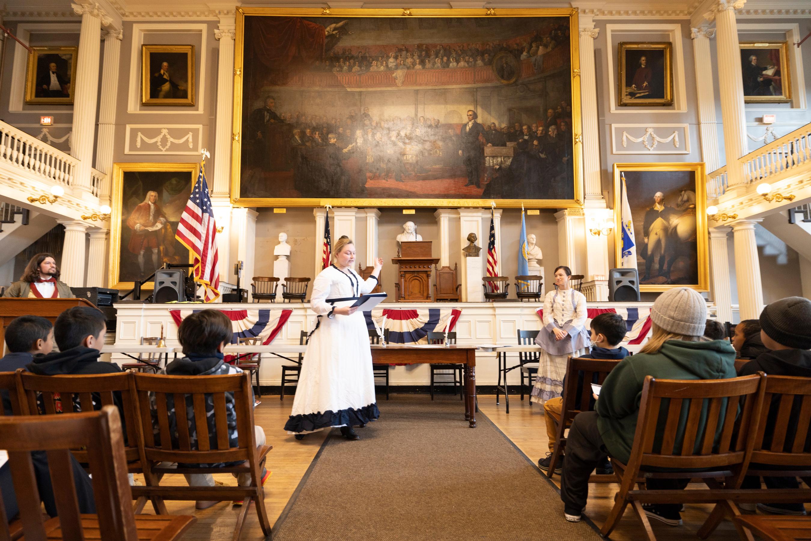 three adults in 1800s clothing stand in front of the stage Faneuil Hall as students look on.