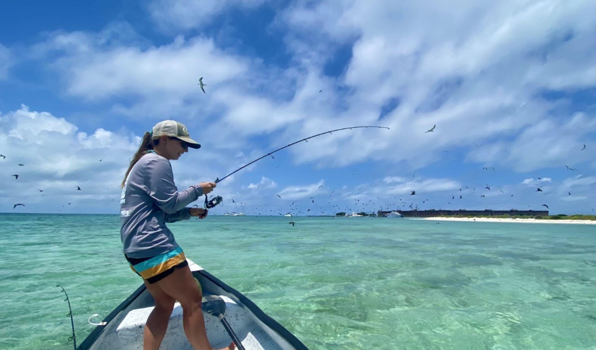 A female angler fights a fish in a small boat atop of crystal clear blue waters.