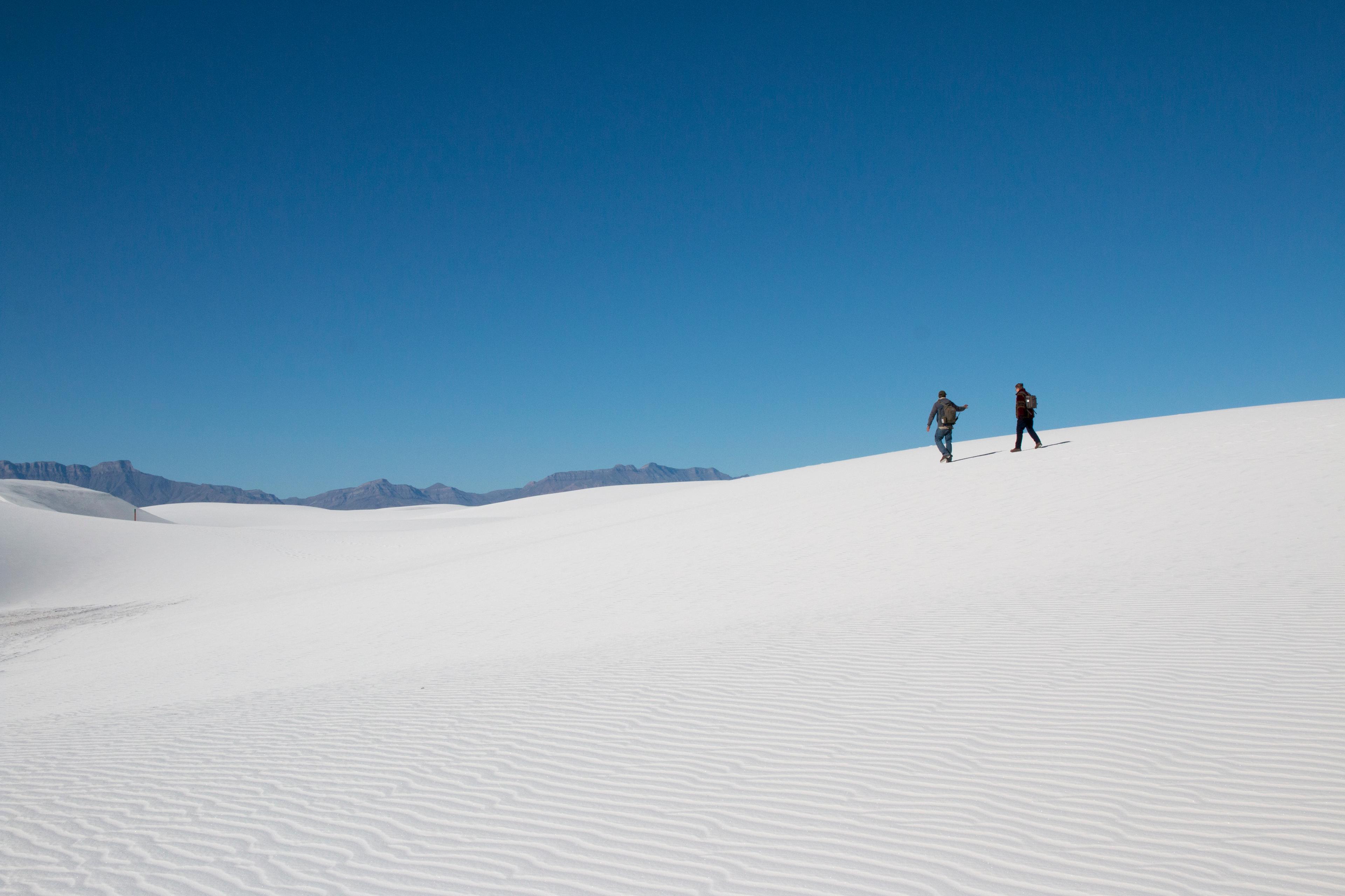 Two people silhouetted on top of a white dune with blue sky overhead