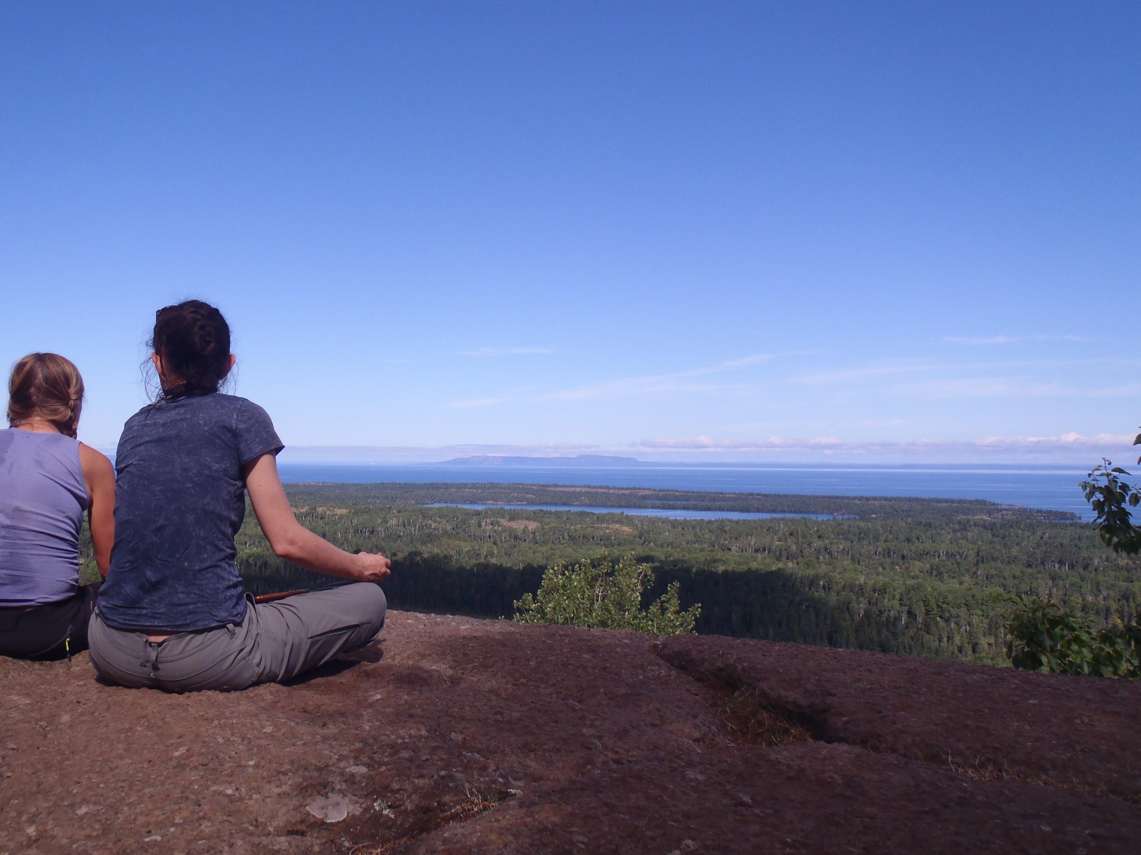 Two people sit on a rocky outcropping over looking a forest and lakes on an island in Lake Superior.