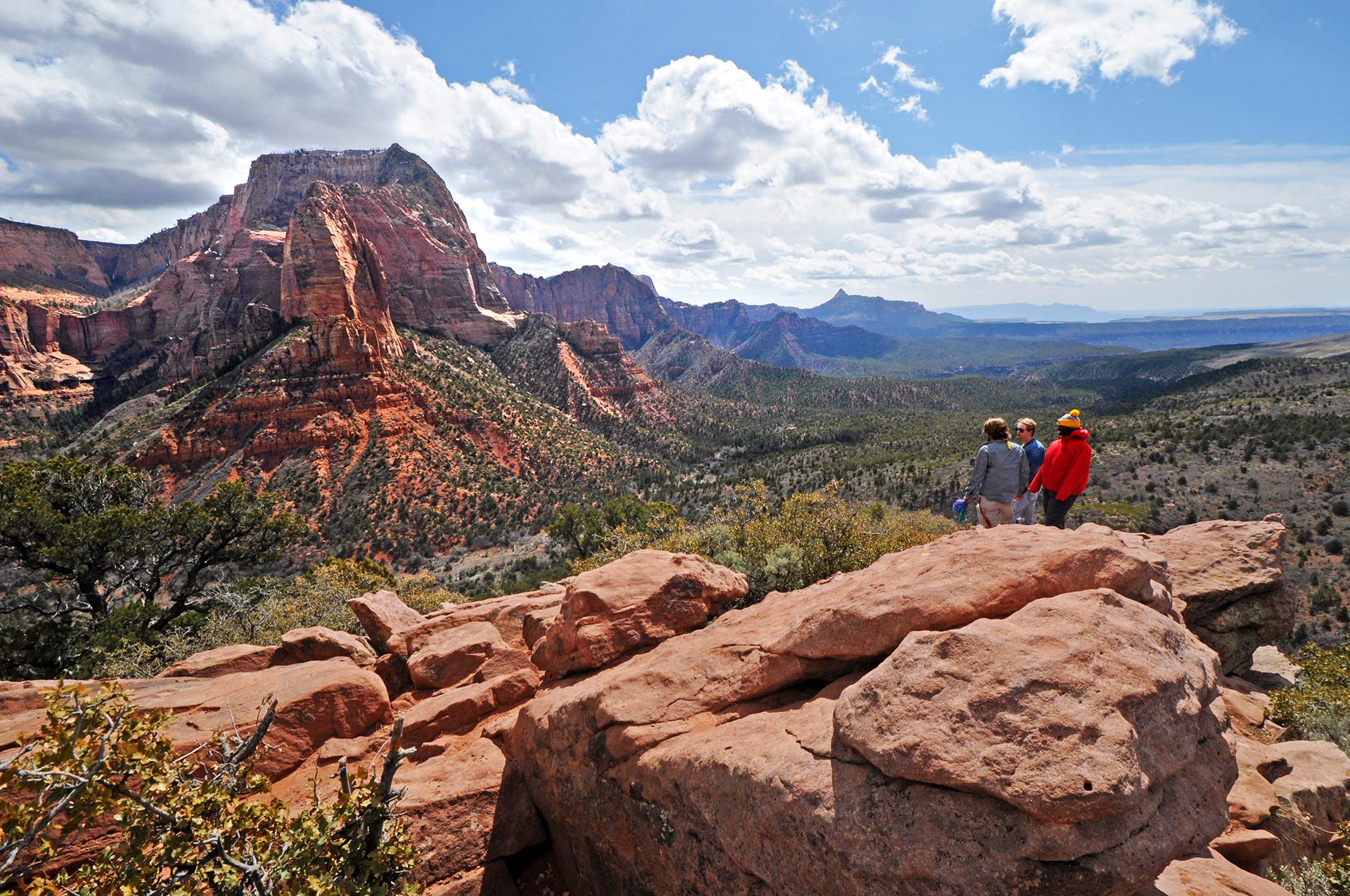 Red sandstone rocks with three hikers standing on them looking out over a valley. Blue, cloudy sky.