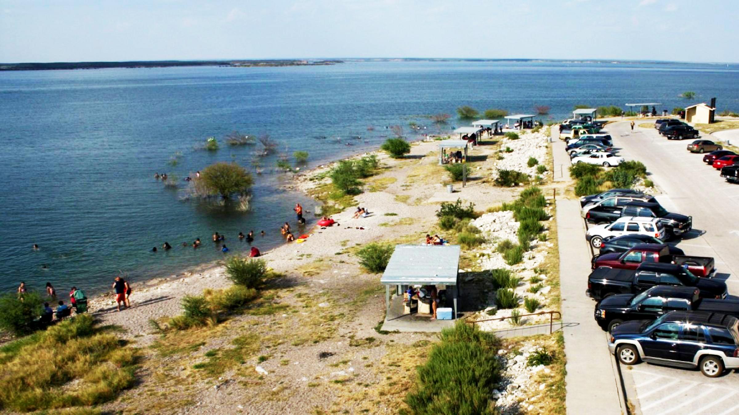 Lines of cars in parking lot with a sheltered picnic tables to left of image and people in lake..