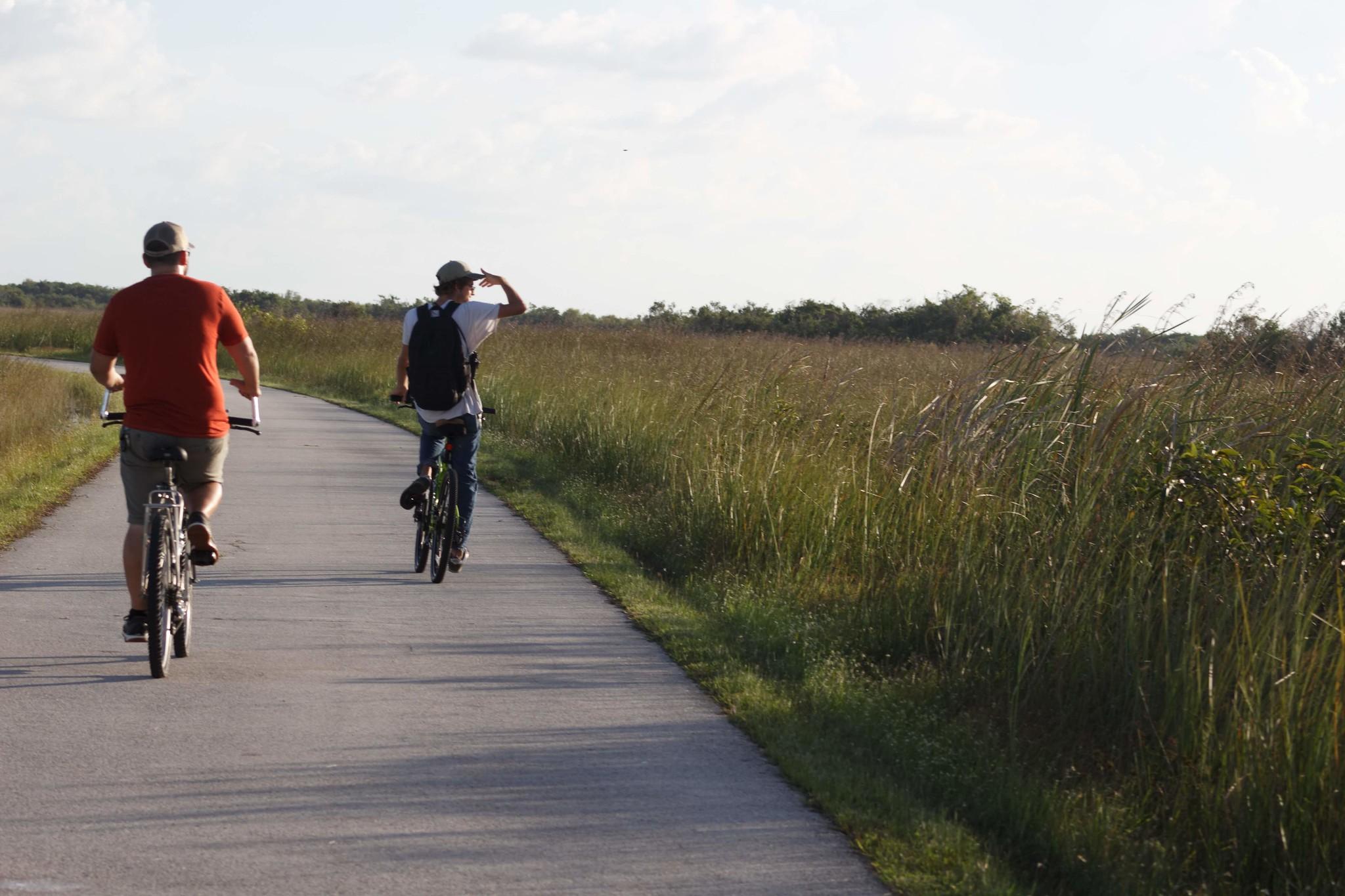 Two bikers peddle along a paved road flanked by open prairie. One bikers stares over the horizon