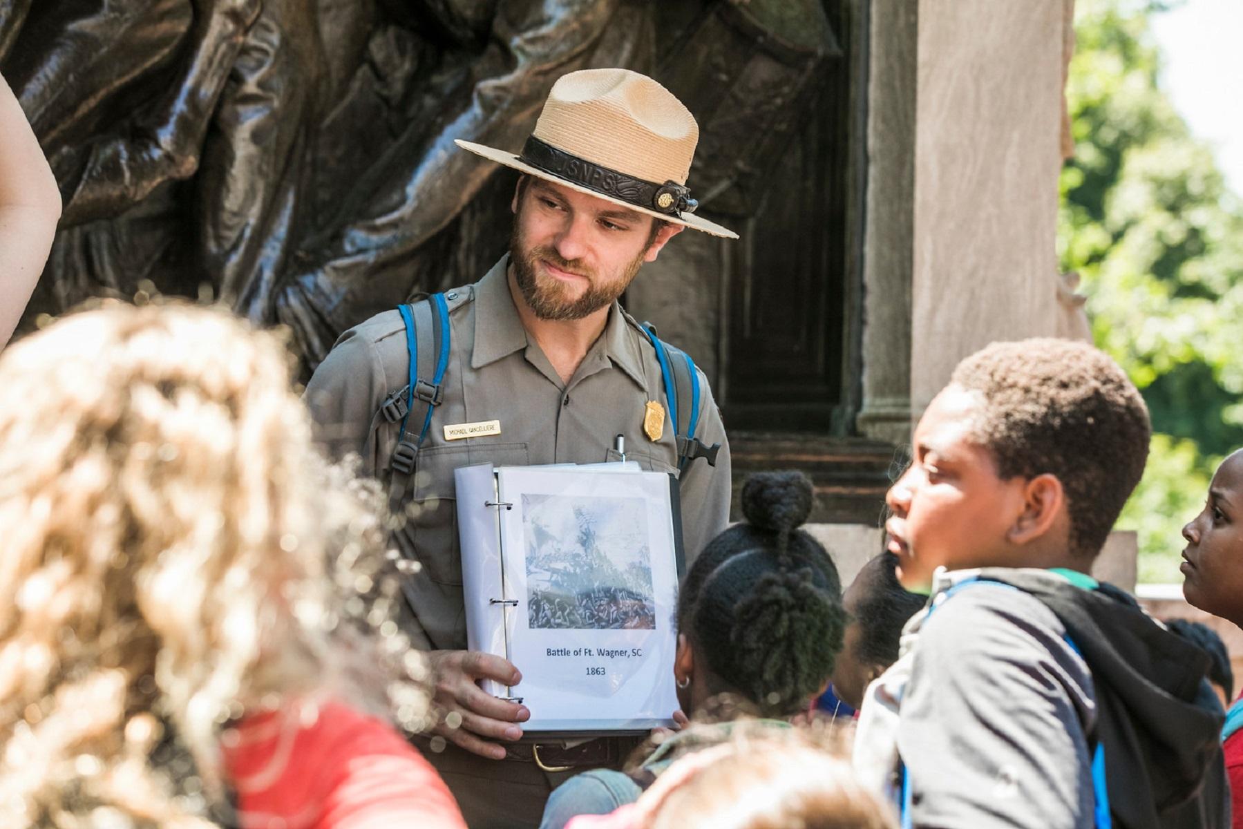 An NPS Ranger speaking to a group of children in front of a monument, holding an image of soldiers.