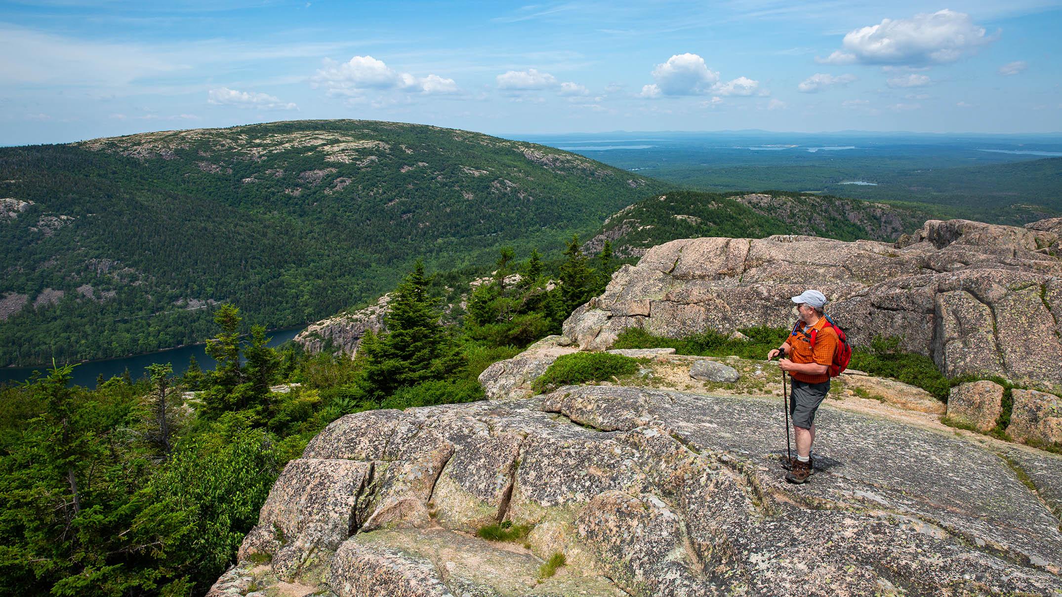 A hiker stands on an exposed granite peak, looking out at mountains rising across the valley.