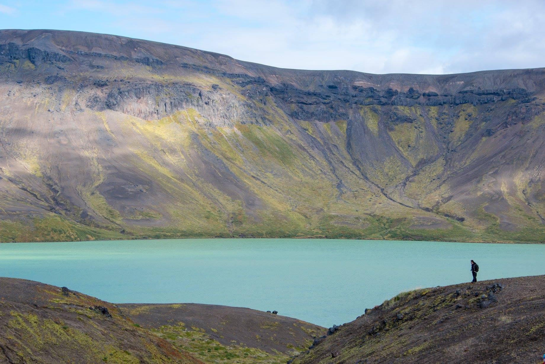 A silhouette of a figure in the distance with a blue lake and caldera rim behind them.