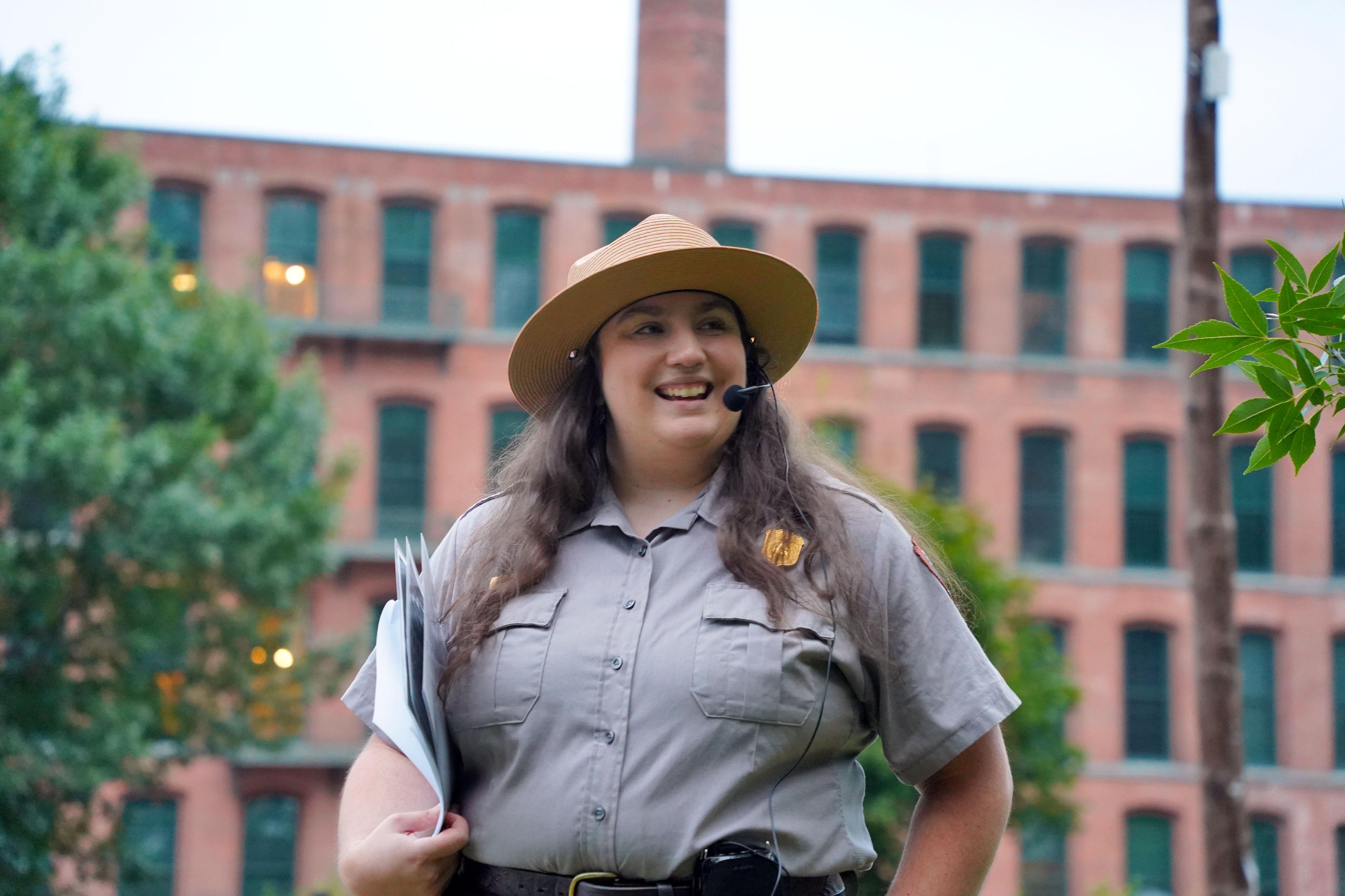 Ranger stands smiling holding papers in front of 4-story brick mill