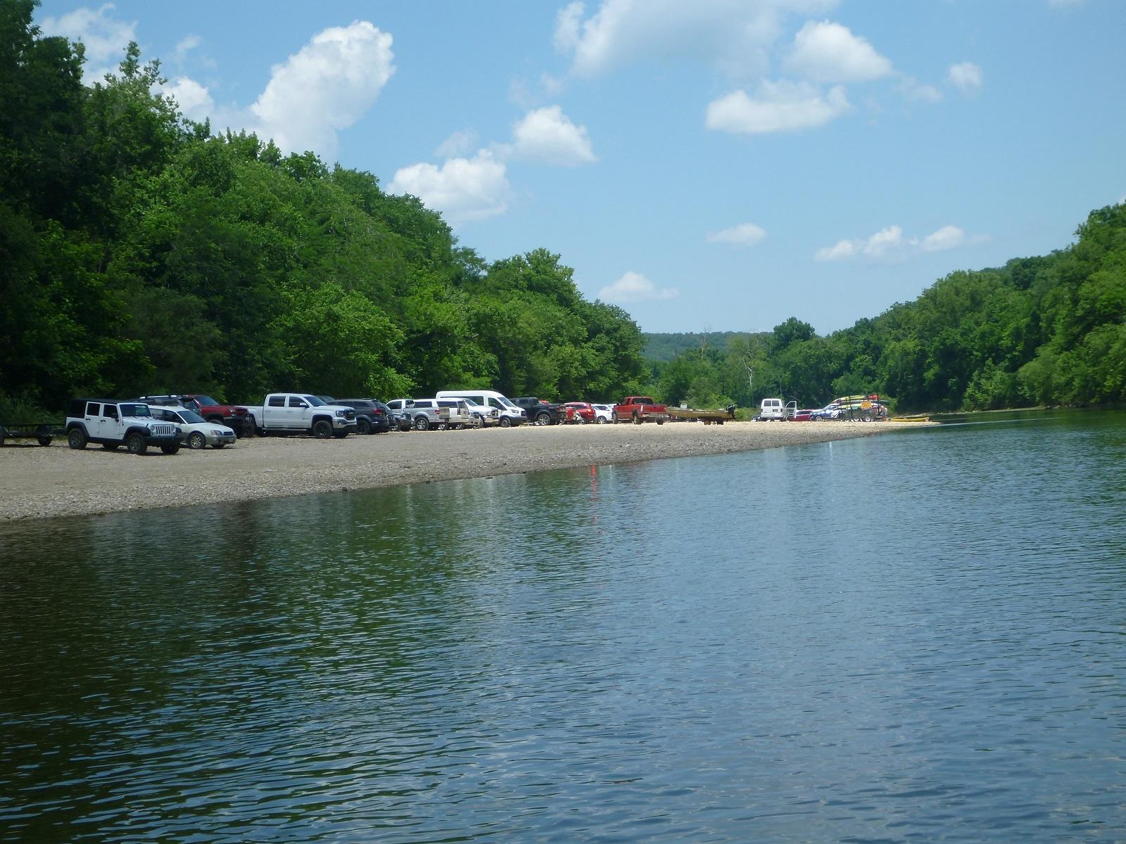 blue river in foreground with many cars parked on tan gravel at center