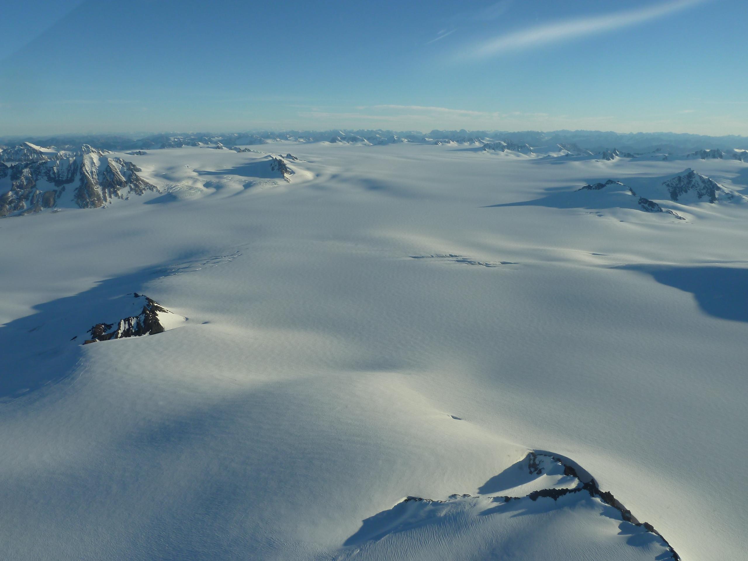 An aerial view of a glacier. Snow covers the land, and mountain tops appear from underneath the ice