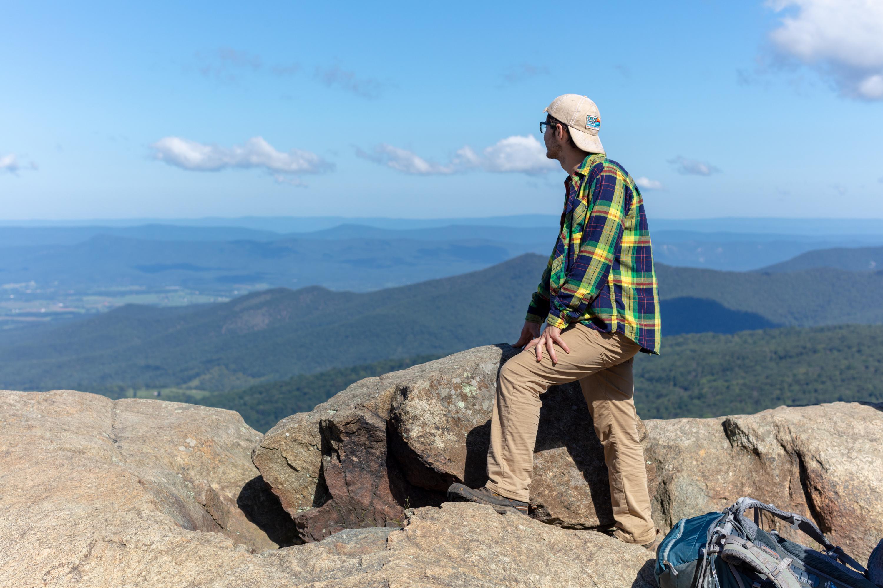 A man in a green shirt and backwards ball cap gazes out over a valley below from a mountain top.