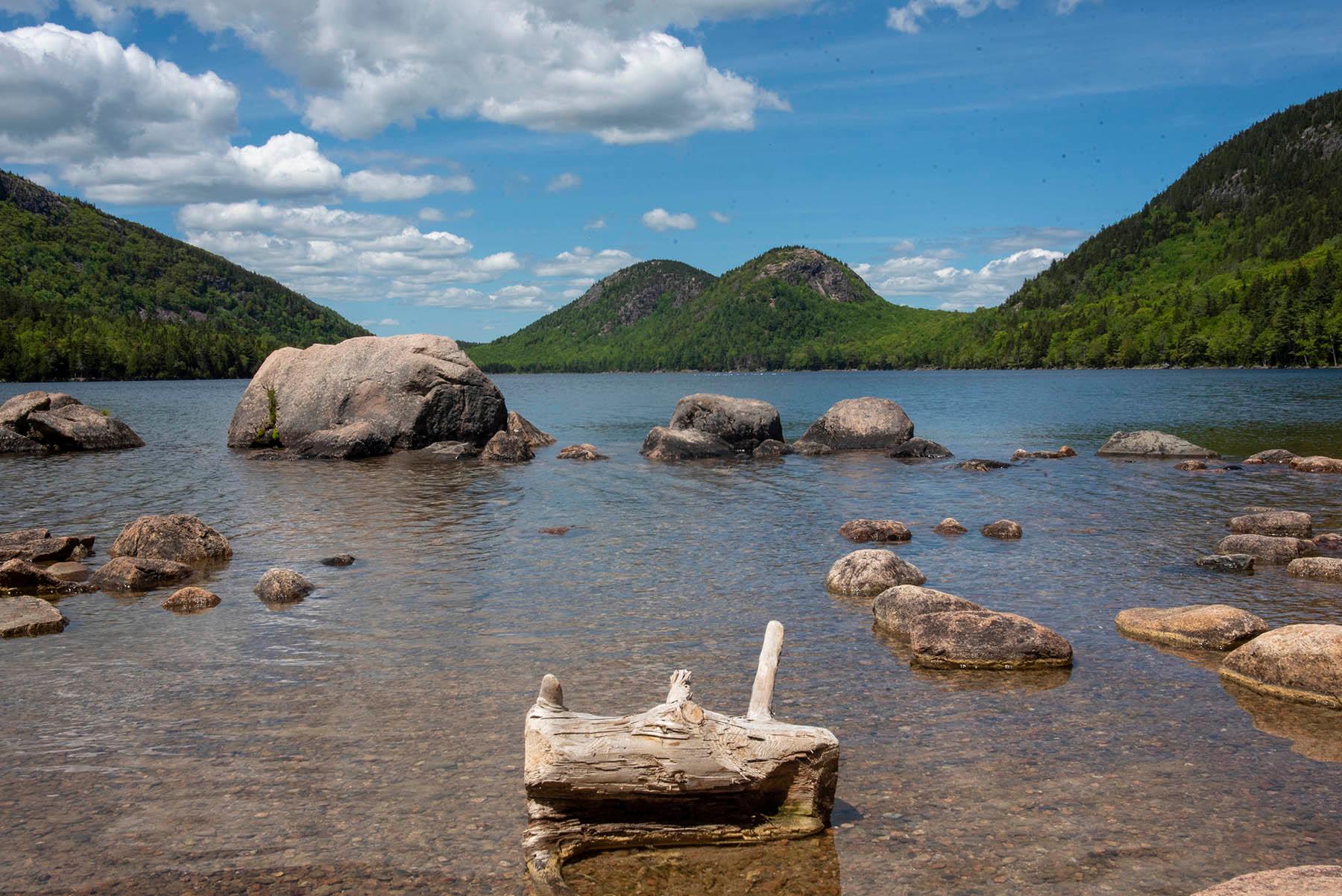 The placid surface Jordan Pond with exposed boulders and a log protruding from the water.