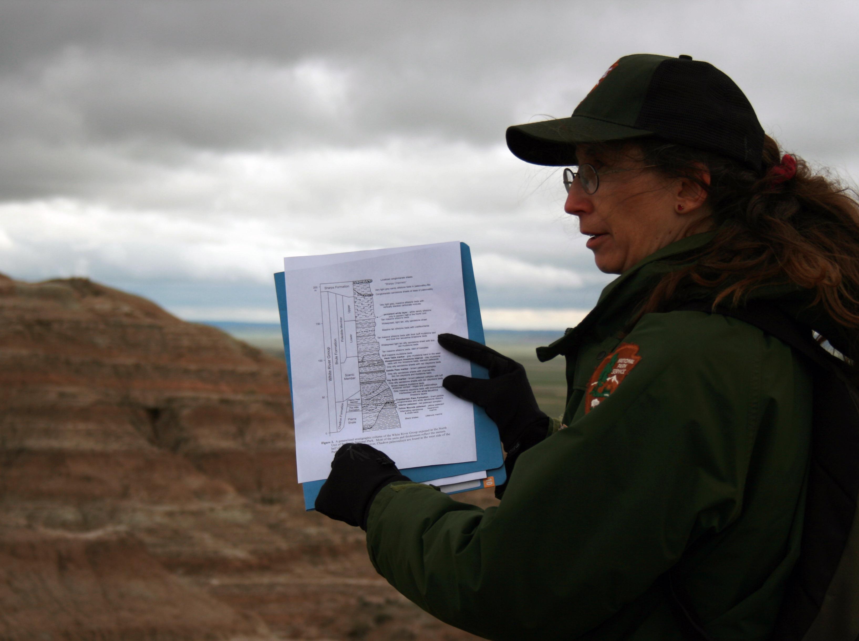 a ranger points at a geologic column on a piece of paper in front of a badlands butte.