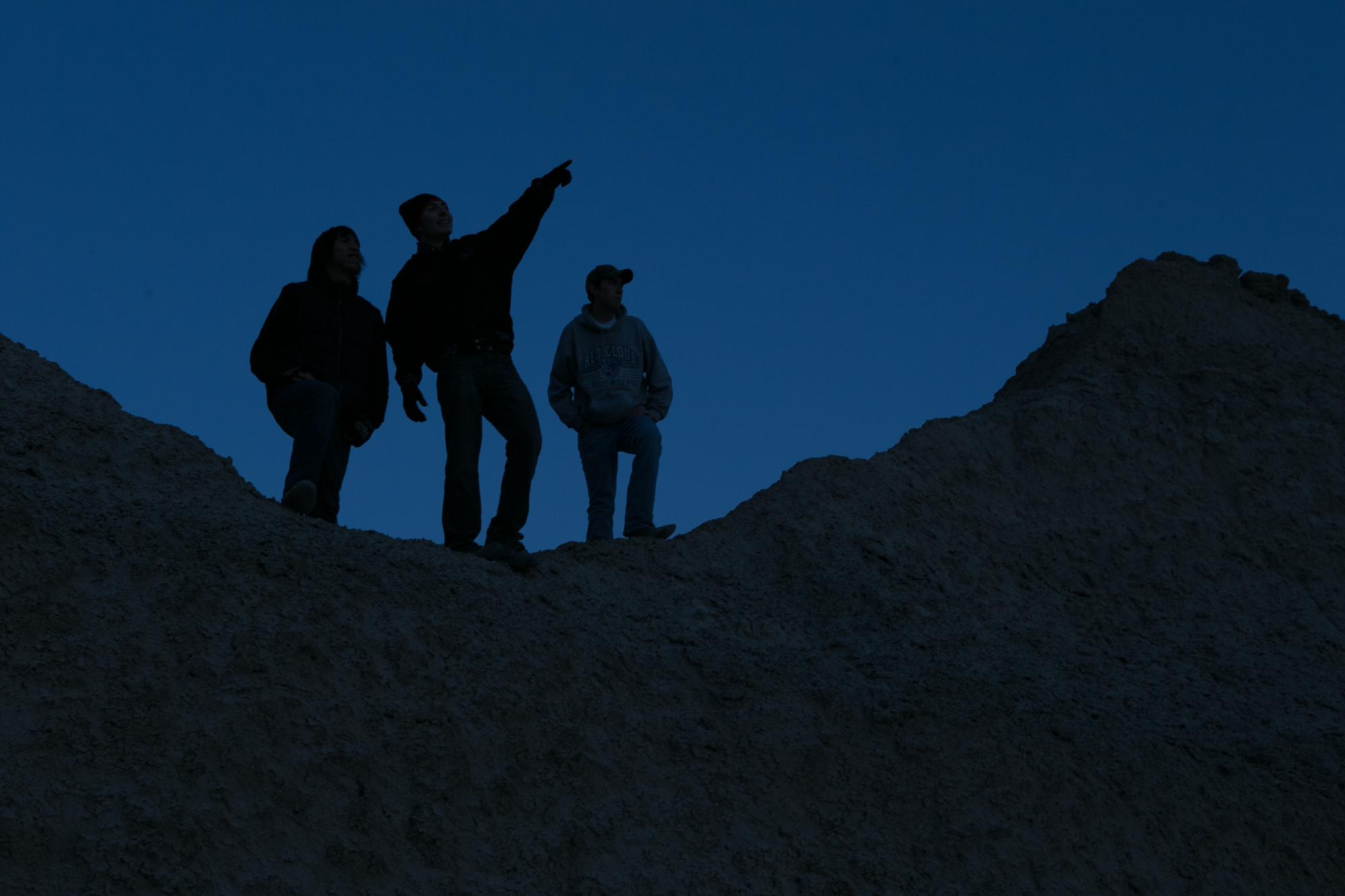 three people stand silhouetted atop a badlands butte, pointing at the night sky.