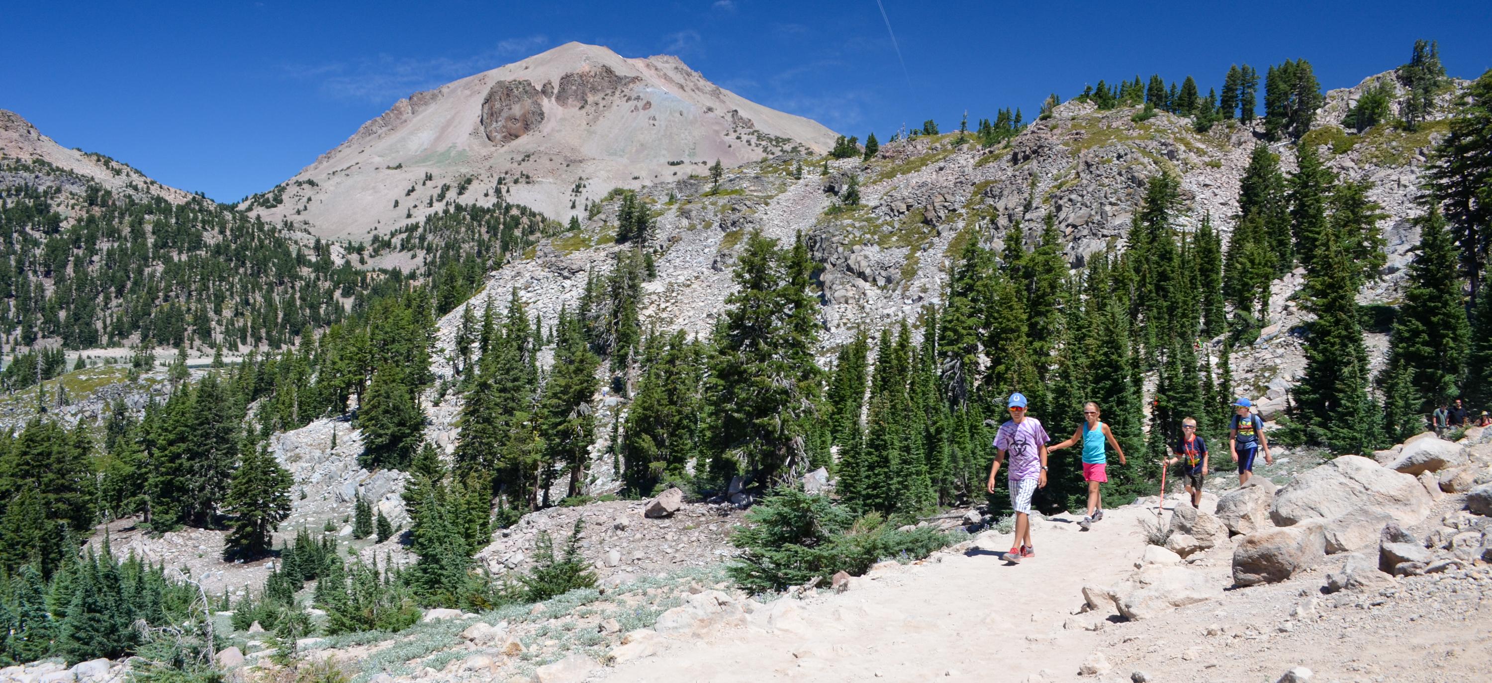 Four young kids hiking on a trail backed by a large volcanic peak and rocky, tree-lined slopes.