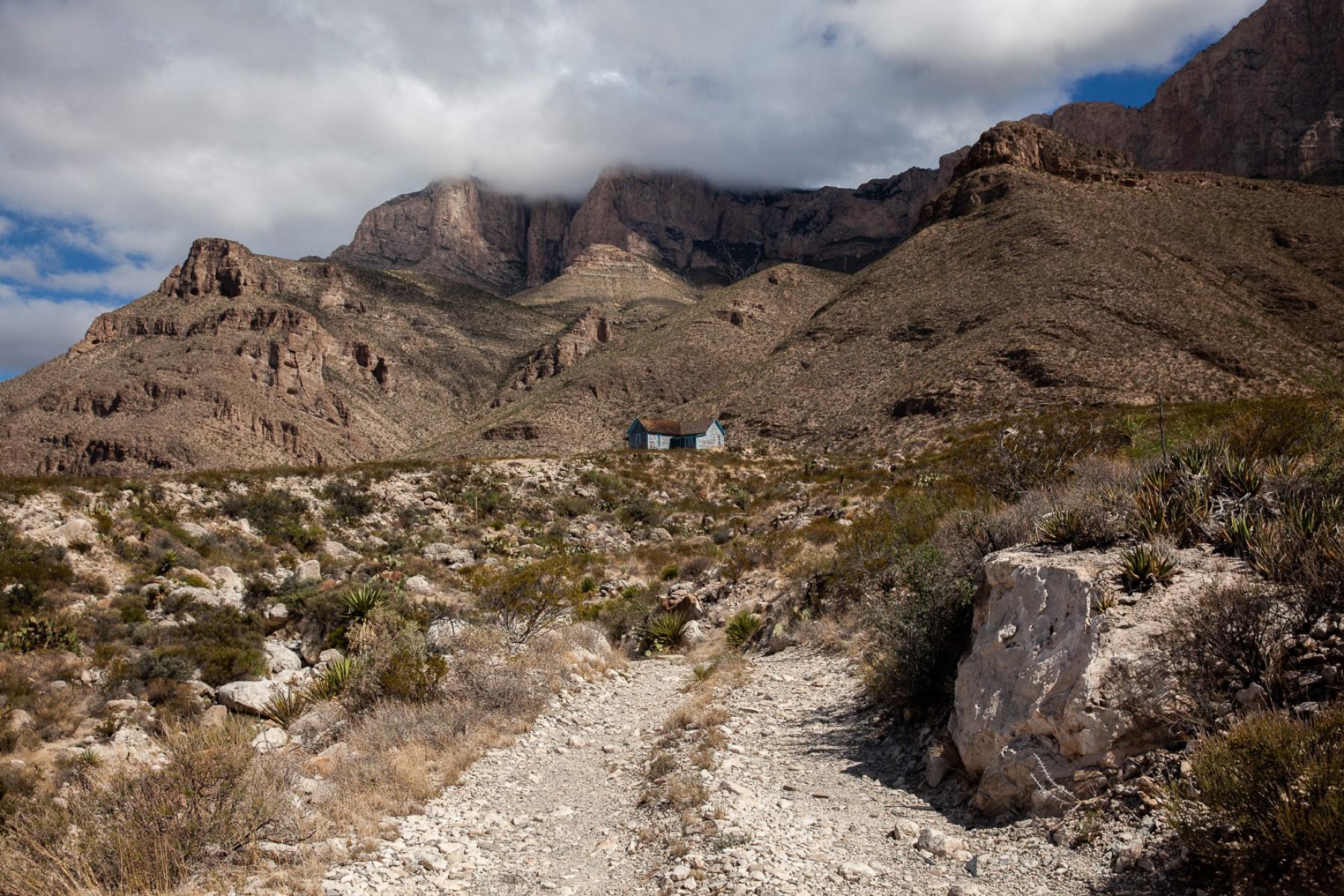 A primitive dirt road leads to a small blue ranch house dwarfed by desert mountains.