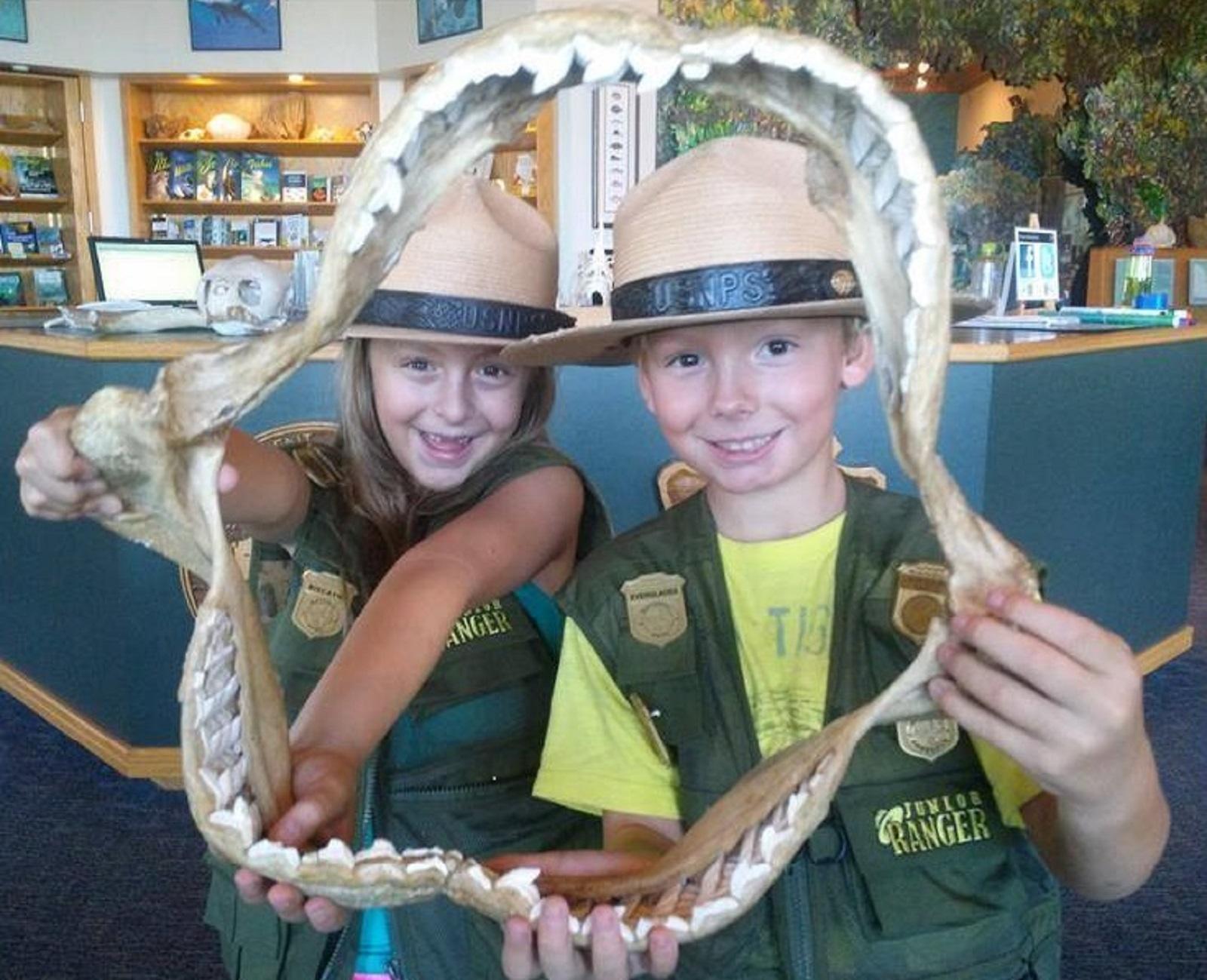 A girl and boy wearing ranger hats and badges peering out from a big shark jaw framing their faces