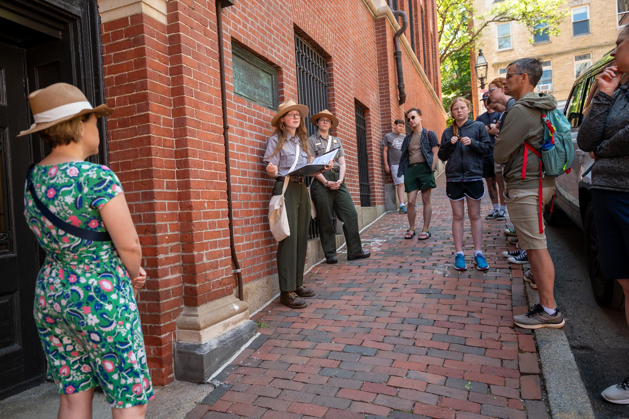 Two park rangers stand in front of a crowd talking next to a brick building