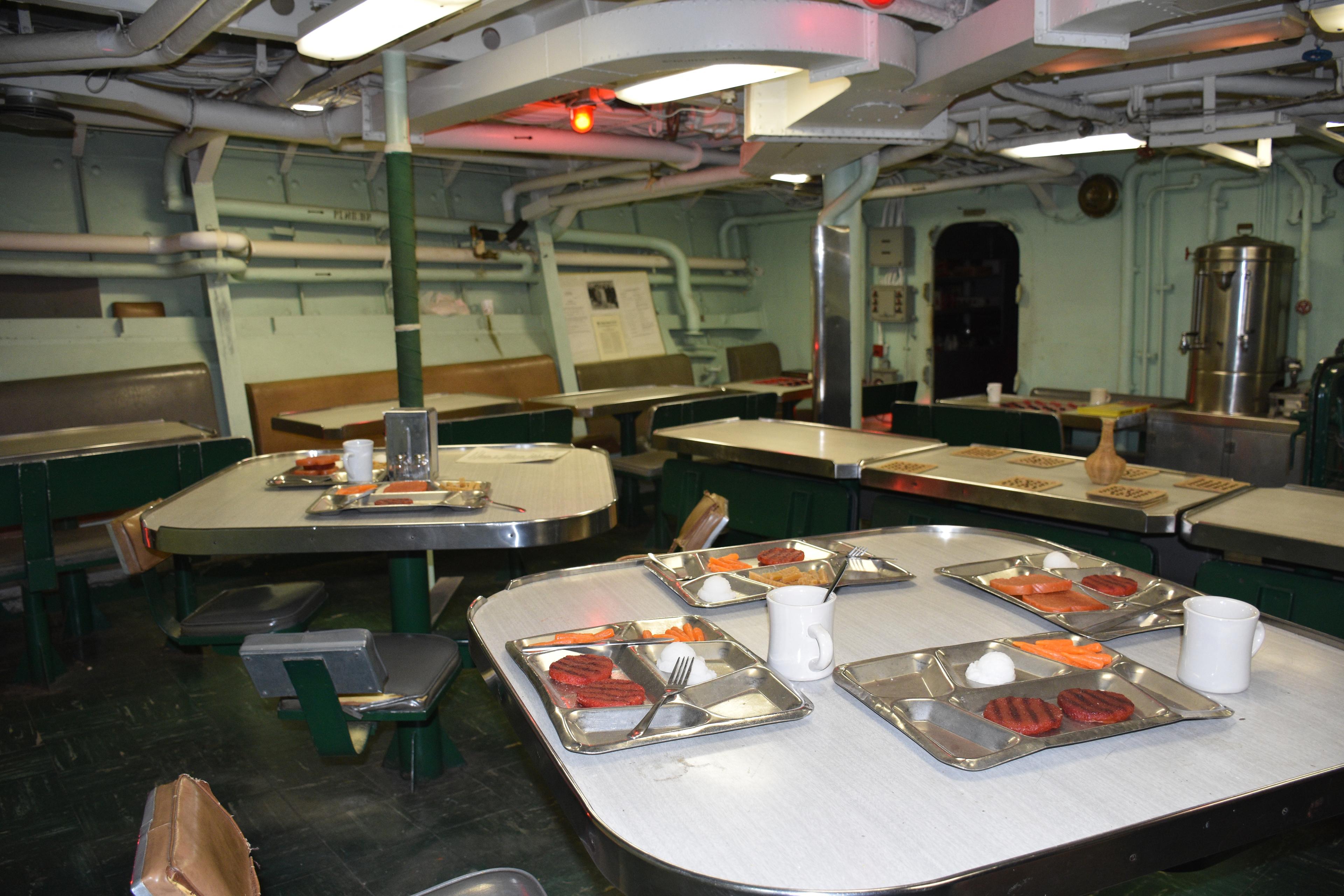 The mess of the USS CASSIN YOUNG which has white square tables with gray food trays on top.