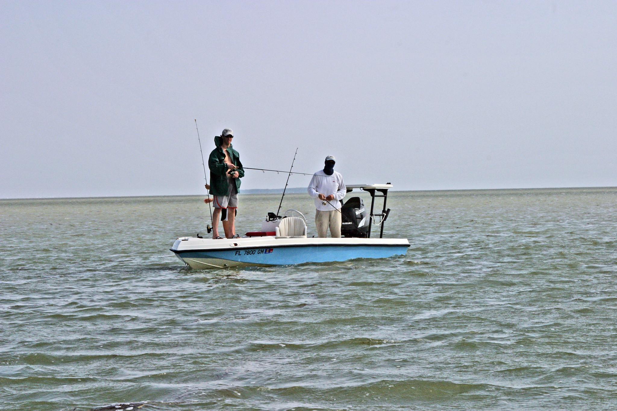 Two anglers stand on a light blue fishing boat in gentle waters. They hold fishing poles in hands