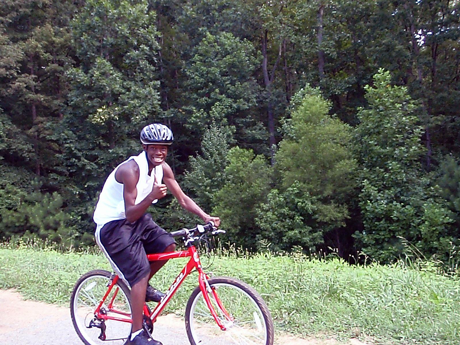 Cyclist giving a thumbs up as he rides on trail with woods in the background.