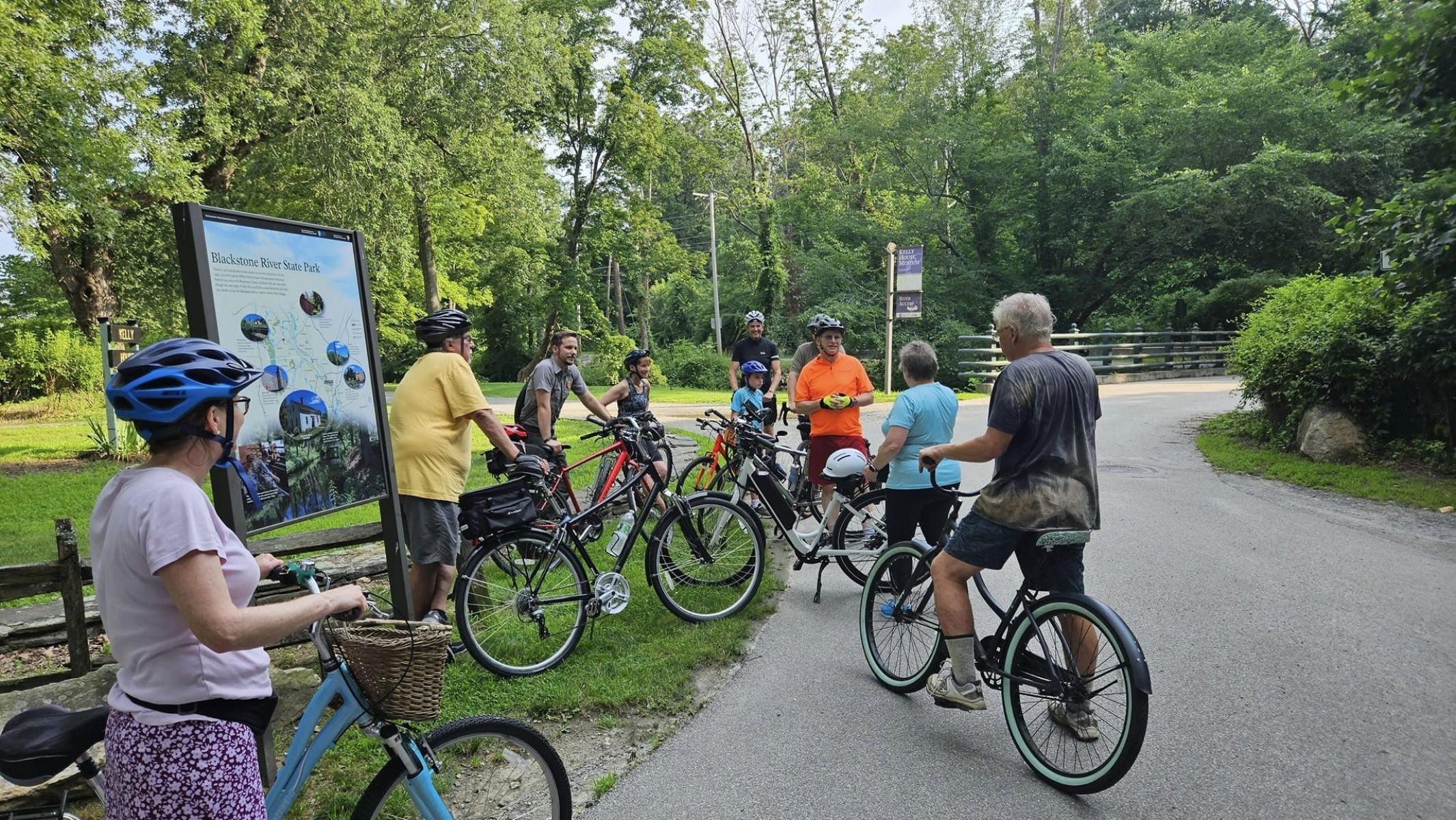 Group of cyclists gather wrong a Ranger and wayside panel for a guided bike ride