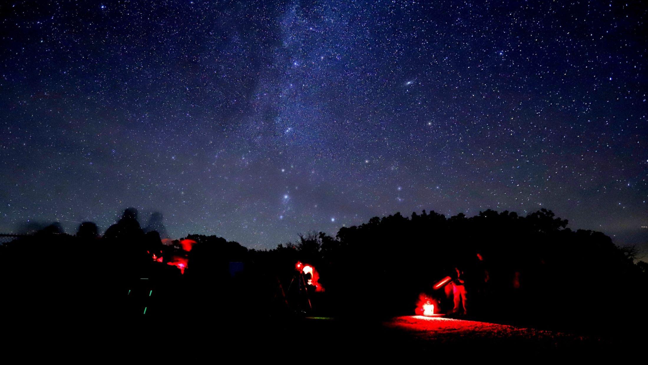 People standing around telescopes with red headlamps underneath a night sky