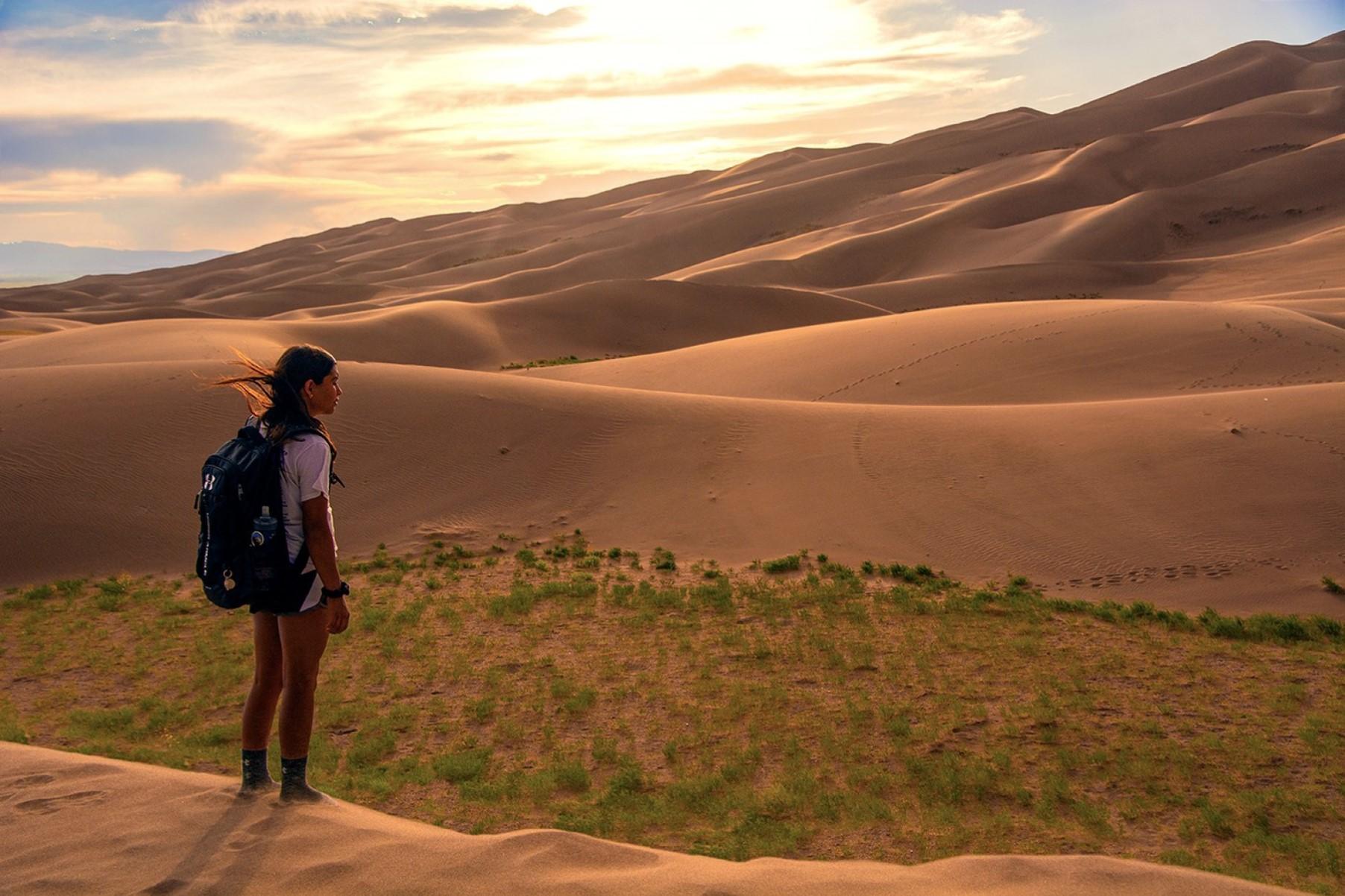 Girl with backpack standing on dunes at sunset