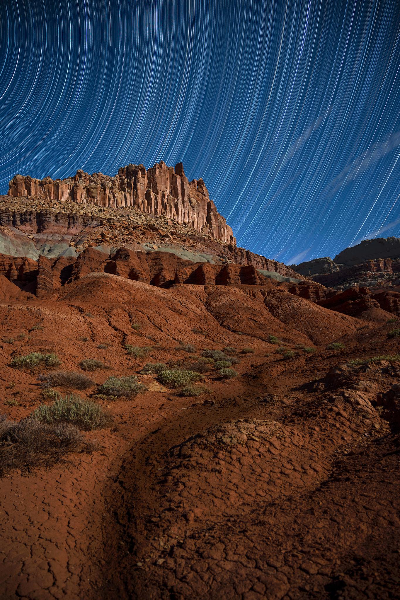 White lines representing circular star trails in dark blue sky, above colorful red cliffs.