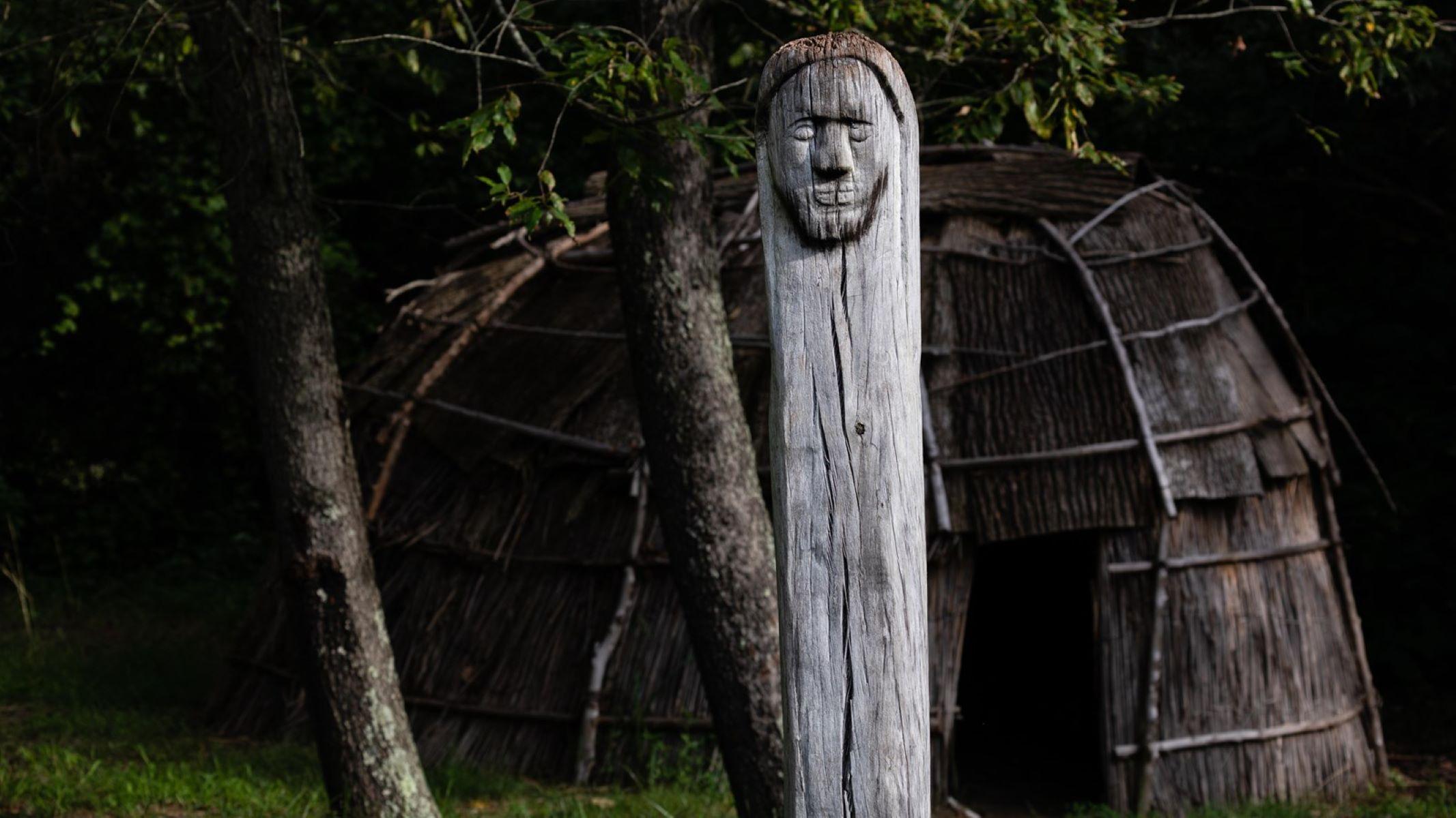 A Native American recreated town has a longhouse structure and sculpture with human face