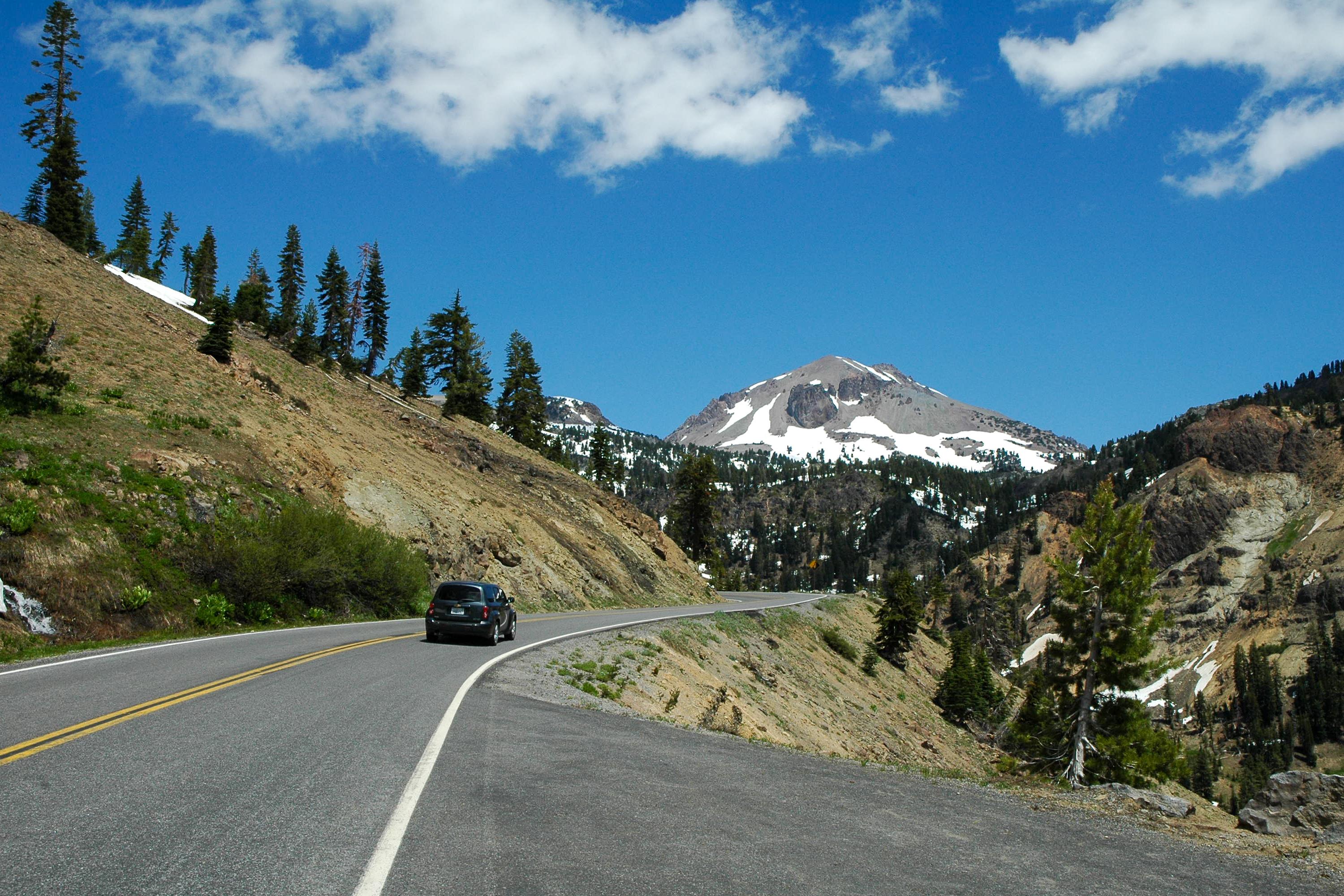 A black vehicle drives on a mountain road toward a large volcanic peak with large patches of snow.