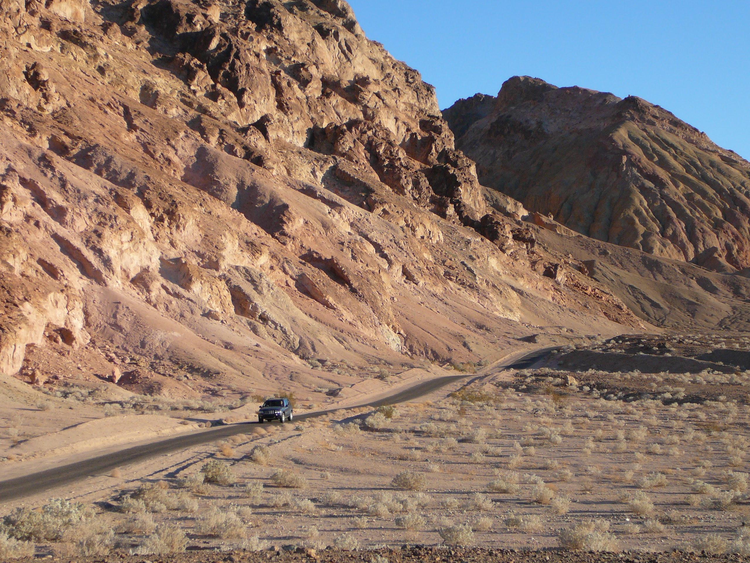 A vehicle drives along a single lane road through colorful, eroded hills.