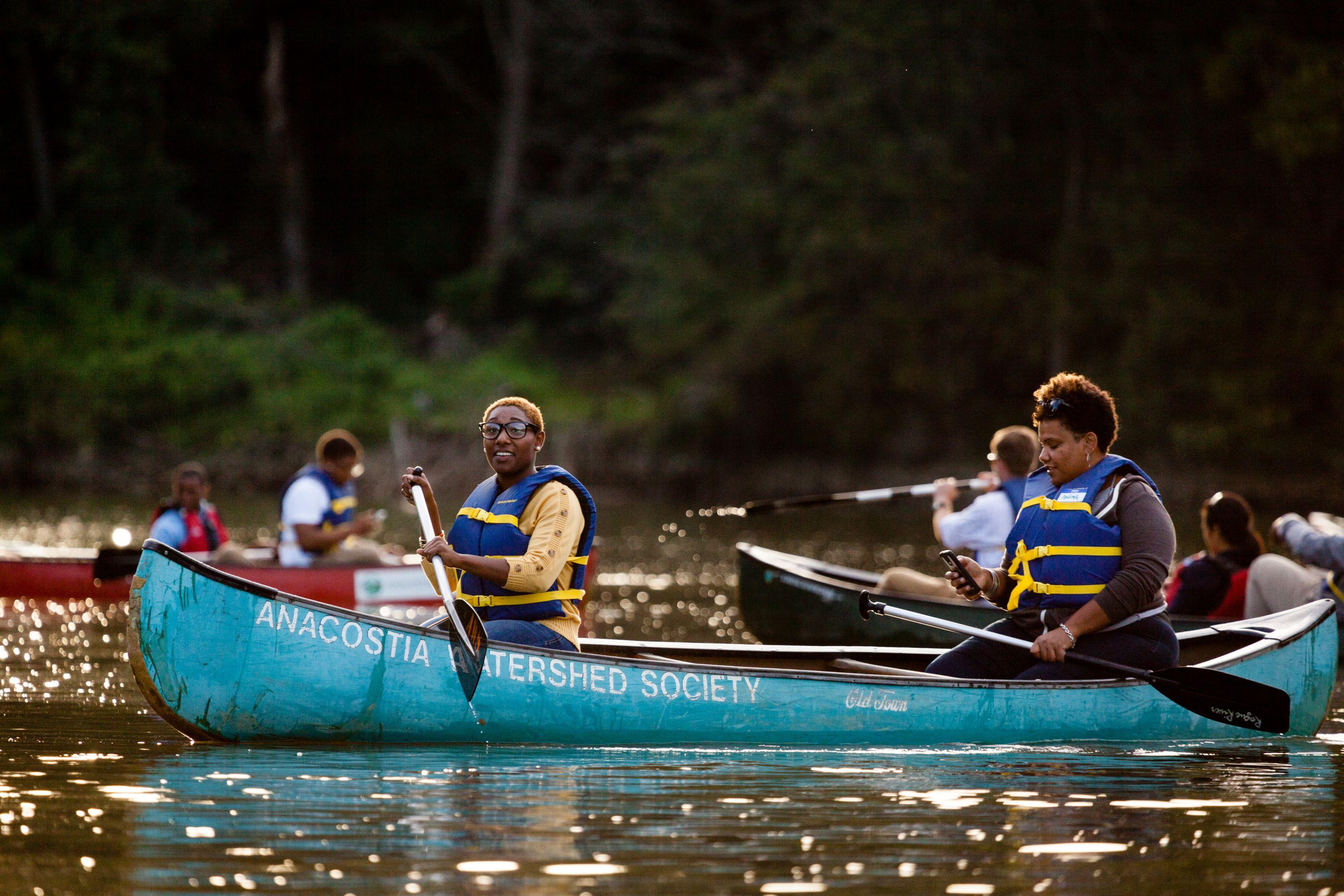 Two people paddle a canoe with two other canoes in the background.