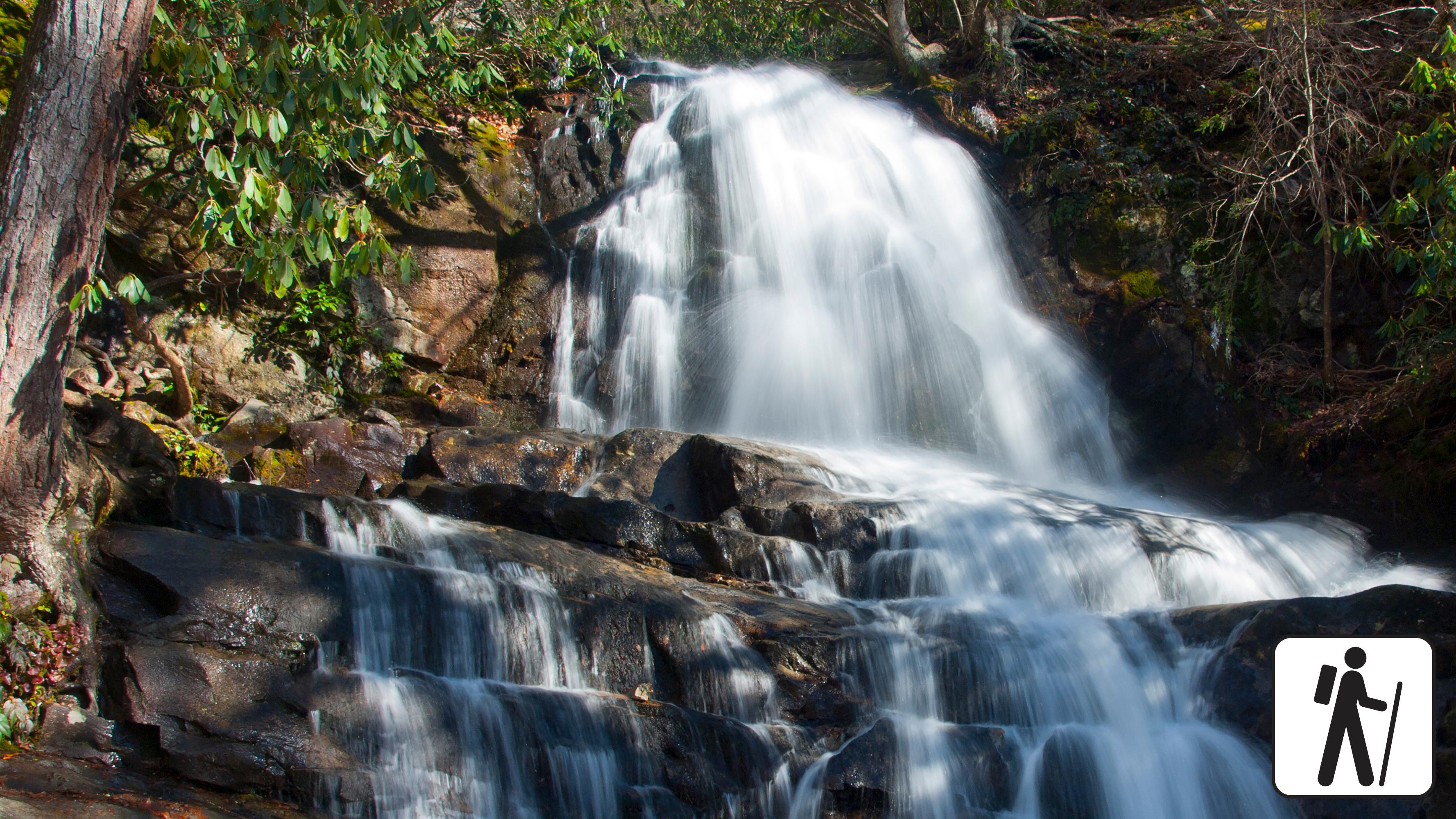 A two-tiered waterfall framed by greenery. Hiker icon in lower right corner.