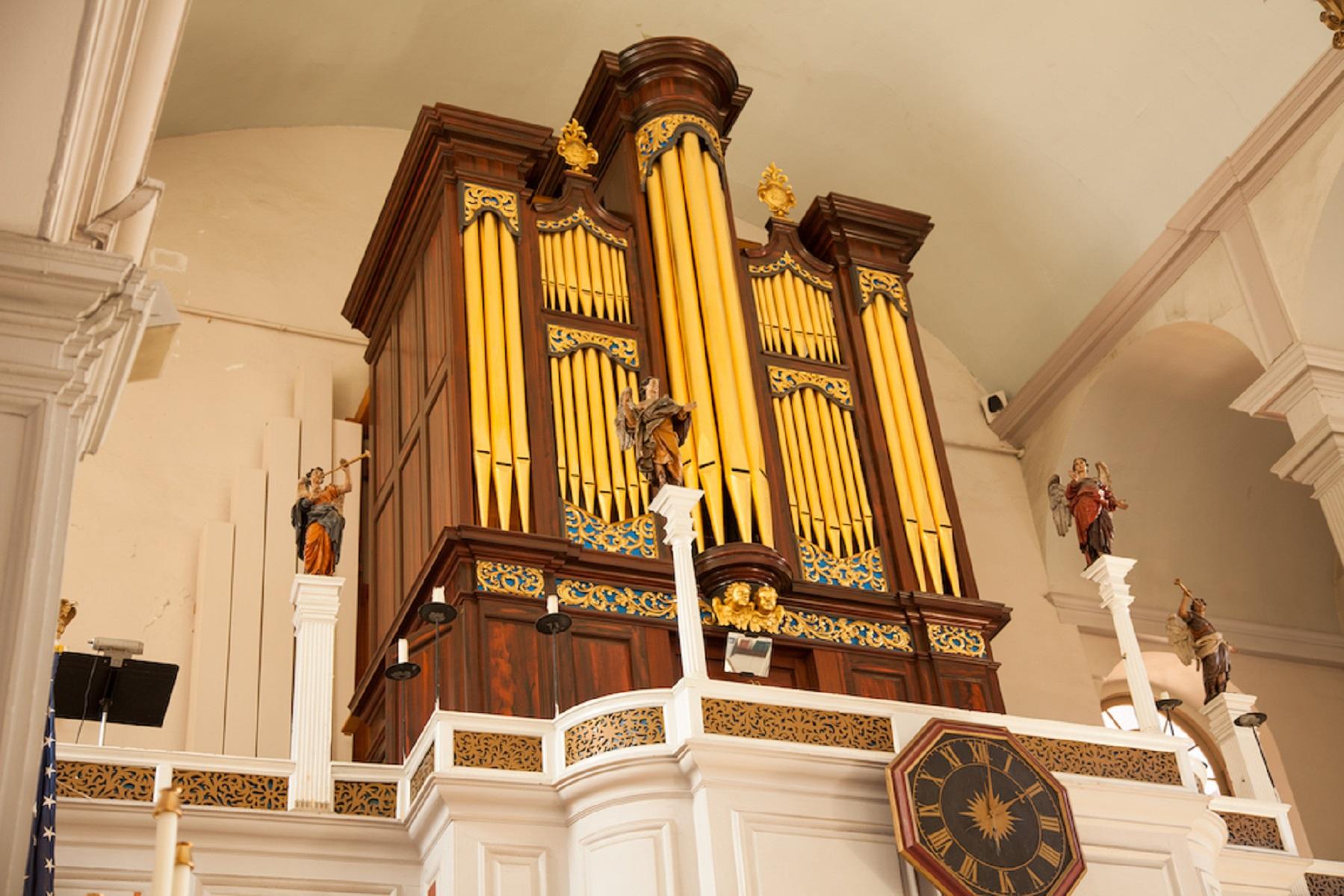 large organ with golden pipes on the second gallery floor of a church. Statues of angels flank it.