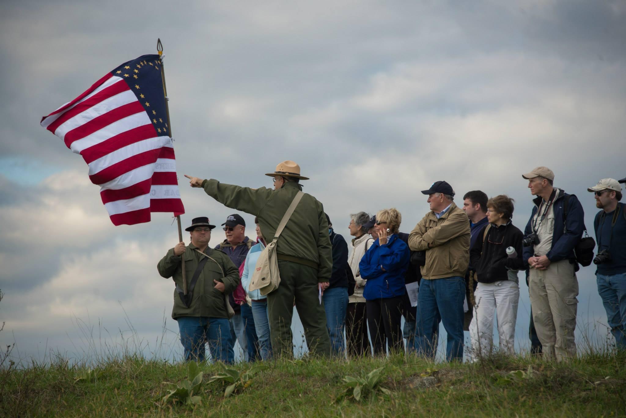 A ranger with visitors on a grass field points past a US flag held by a Civil War reenactor.